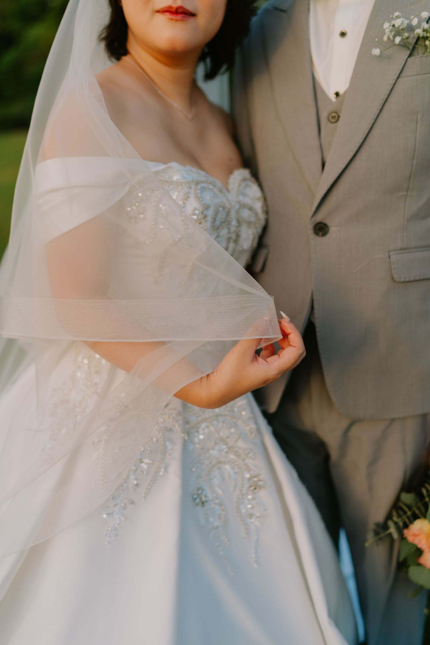 Close-up of bride in beaded off-shoulder wedding gown holding veil beside groom in grey suit.