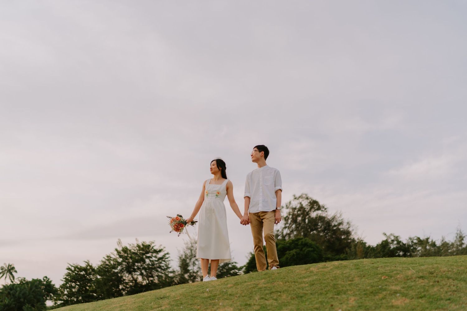 A couple walks hand in hand on a grassy hill under a cloudy sky, the woman holding a bouquet.