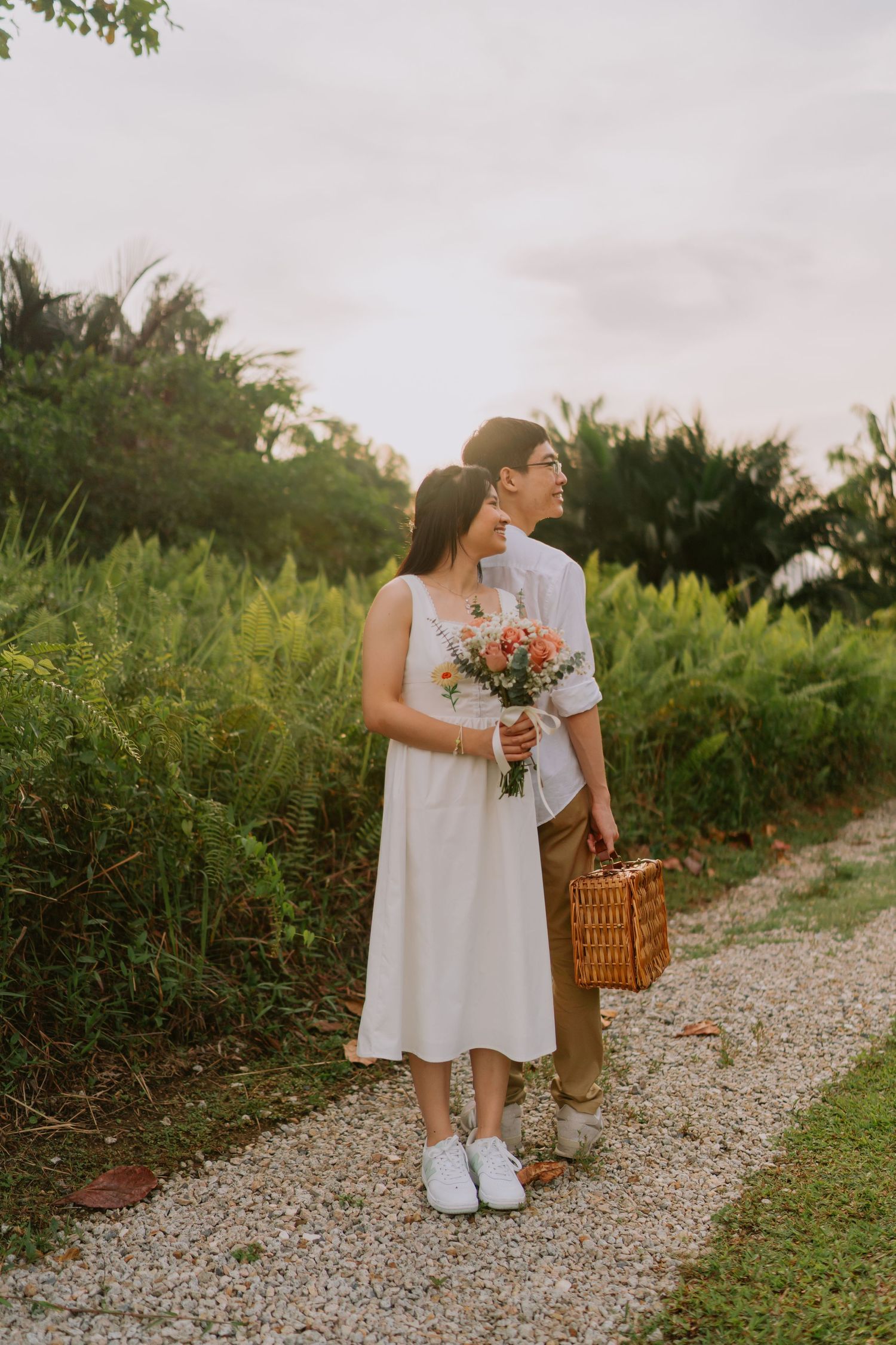 Couple embracing on a gravel garden path at sunset, holding bouquet and wicker picnic basket.