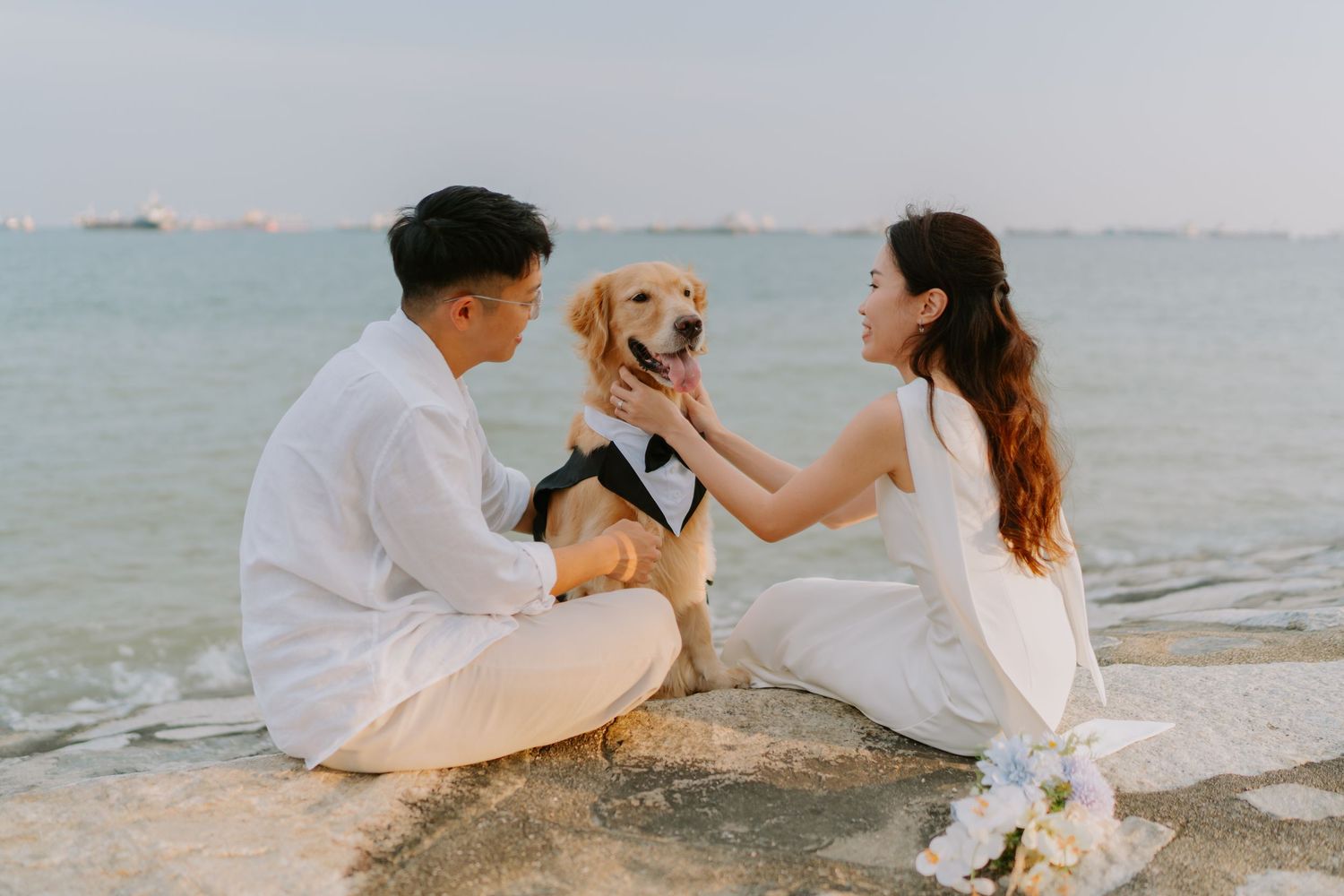 Couple in white outfits adjusting tuxedo on golden retriever while sitting by the sea.