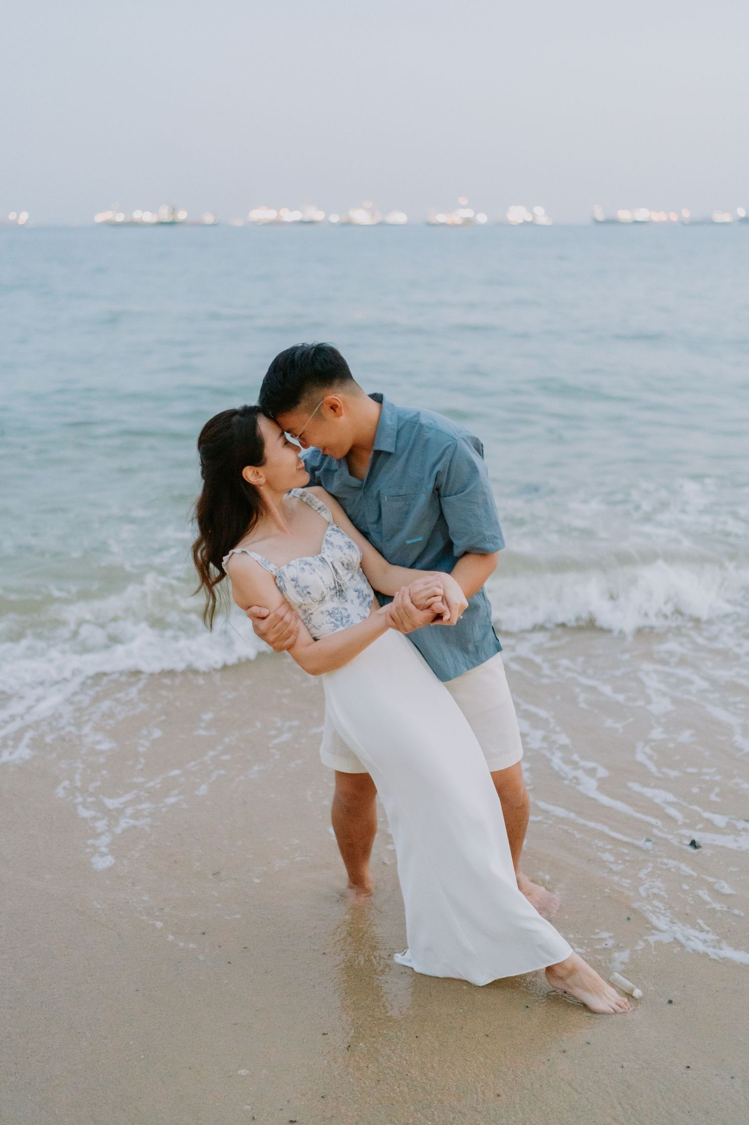 Couple sharing a romantic kiss on the beach at dusk, waves surrounding their feet.