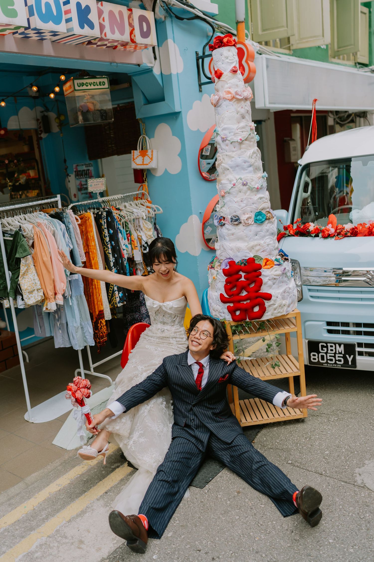 Bride and groom posing playfully outside a vintage shop with a decorative wedding cake sculpture and white van.