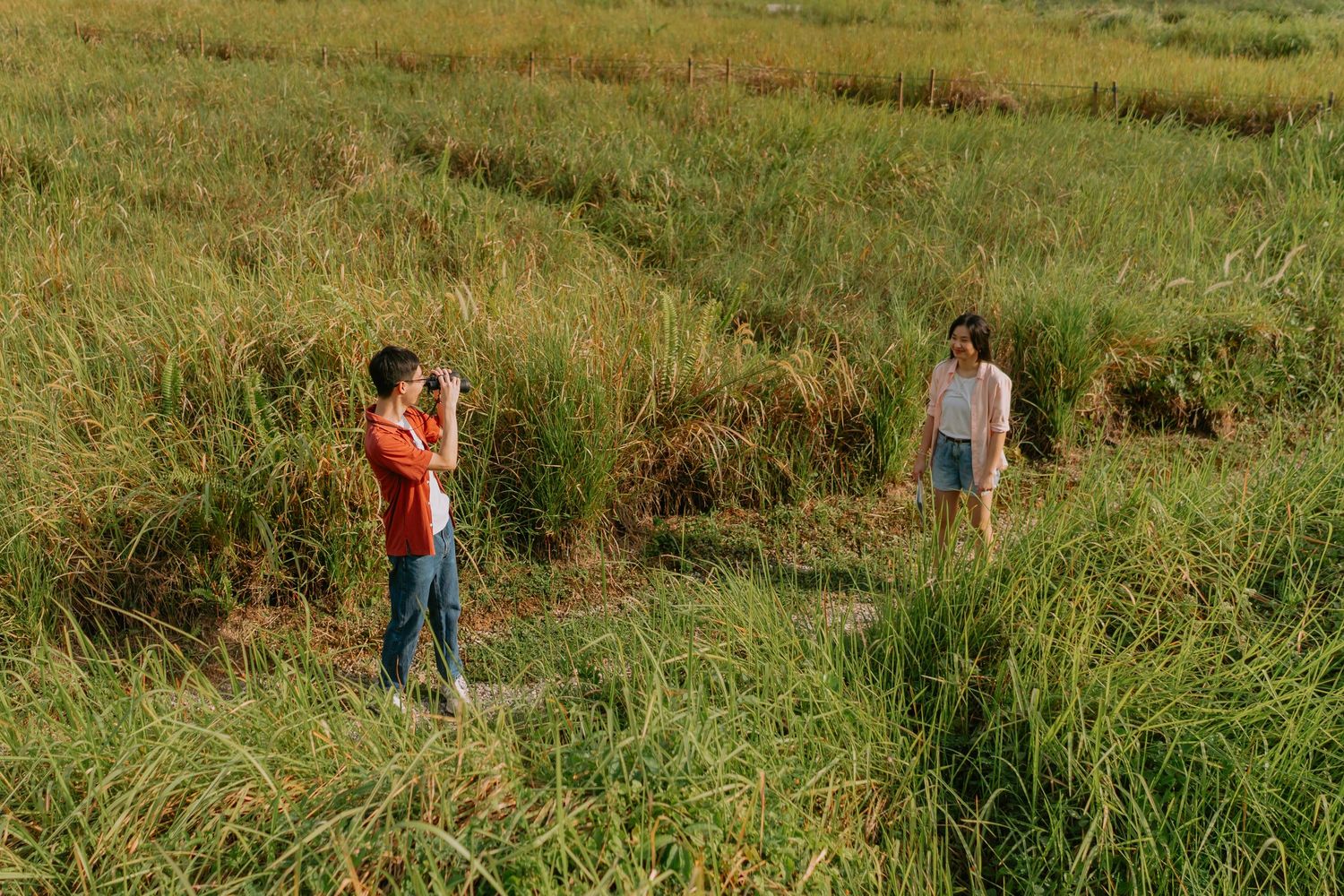 A man photographs a woman standing in a golden grass field during sunset.