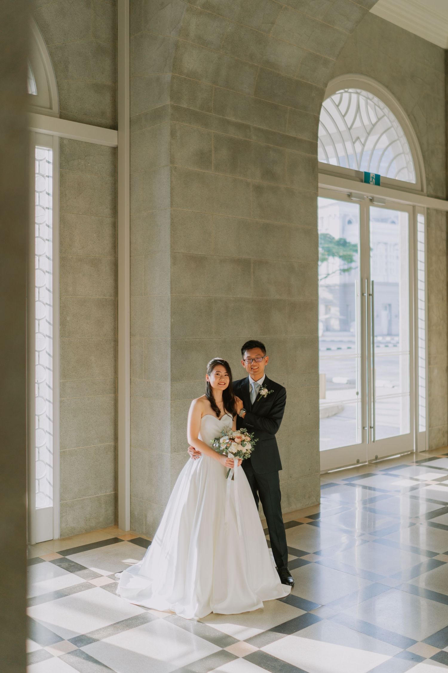 Bride and groom pose together in white marble hall with checkered floor and arched glass doors.