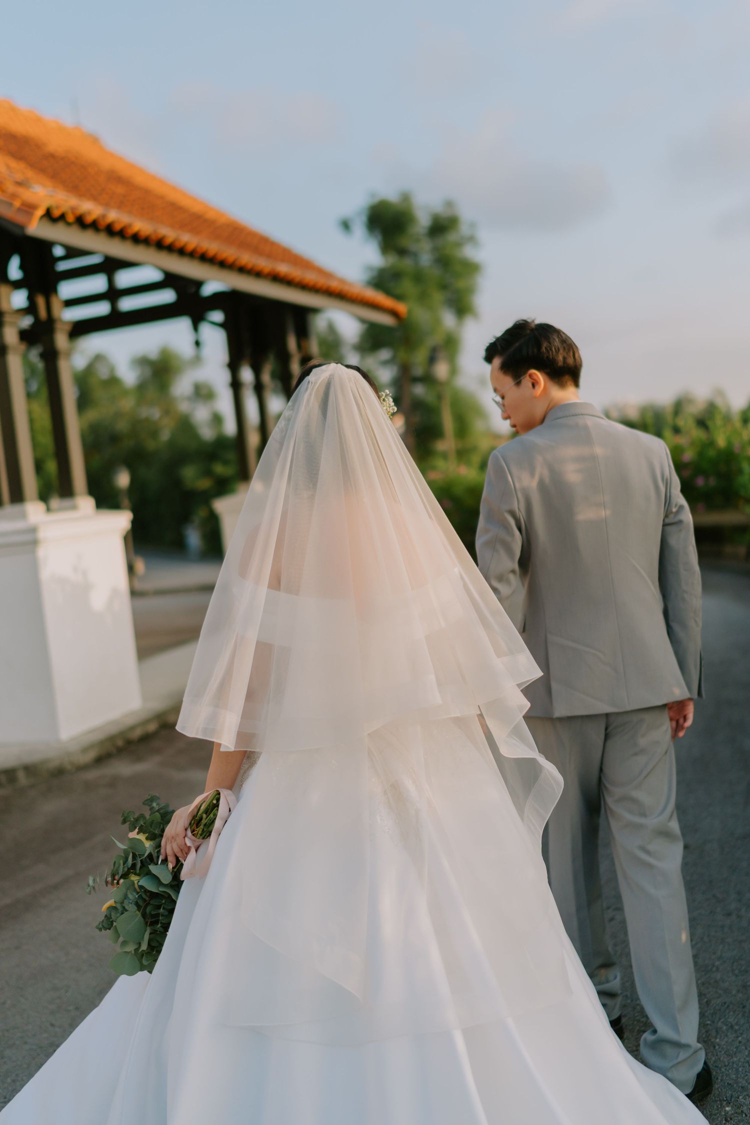Wedding couple walks away from camera, bride in layered veil and white gown, groom in gray suit.