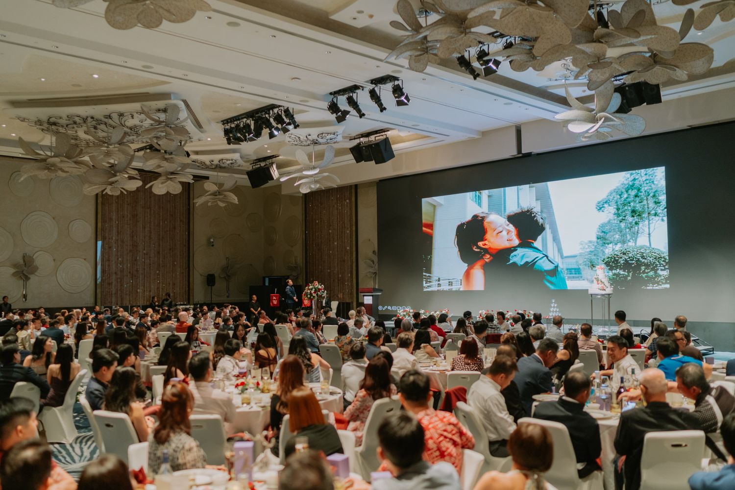 Elegant wedding banquet hall with guests seated at round tables watching a romantic video on a large screen.