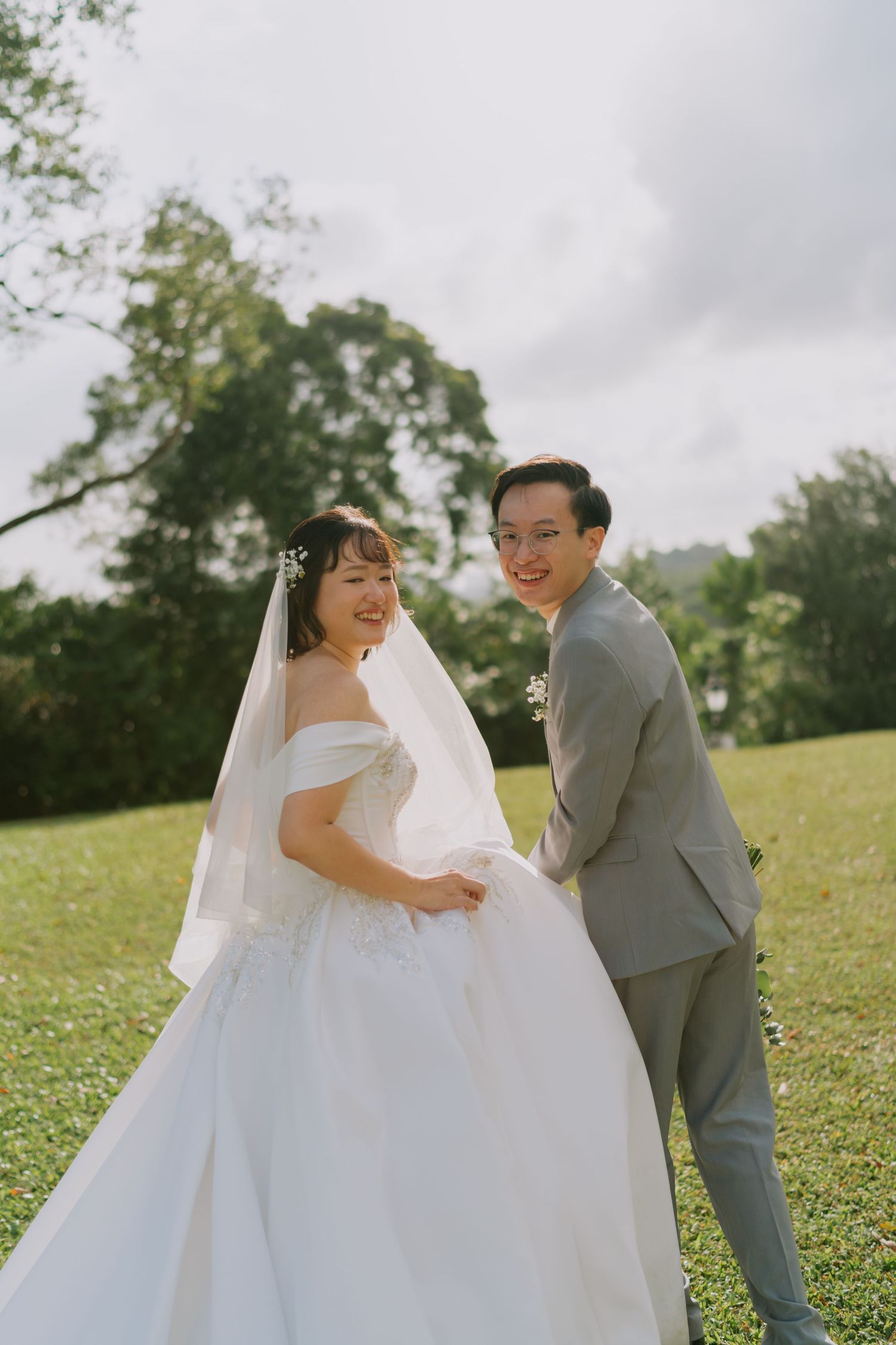 Laughing bride and groom pose together on a green lawn with tall trees in the background.