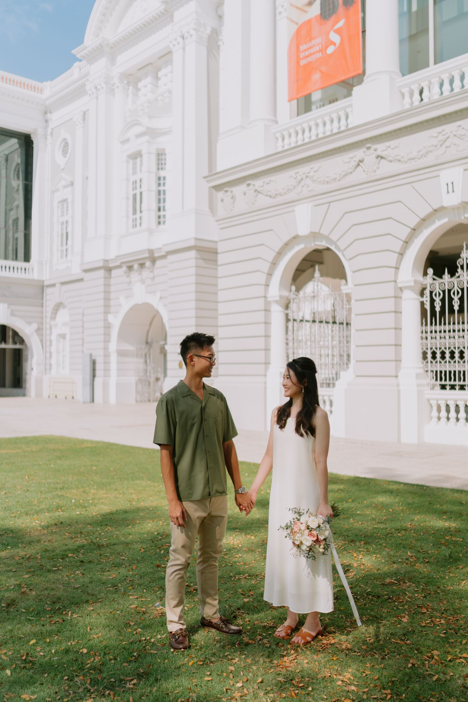 Couple in casual attire hold hands on green lawn before white colonial Singapore building.