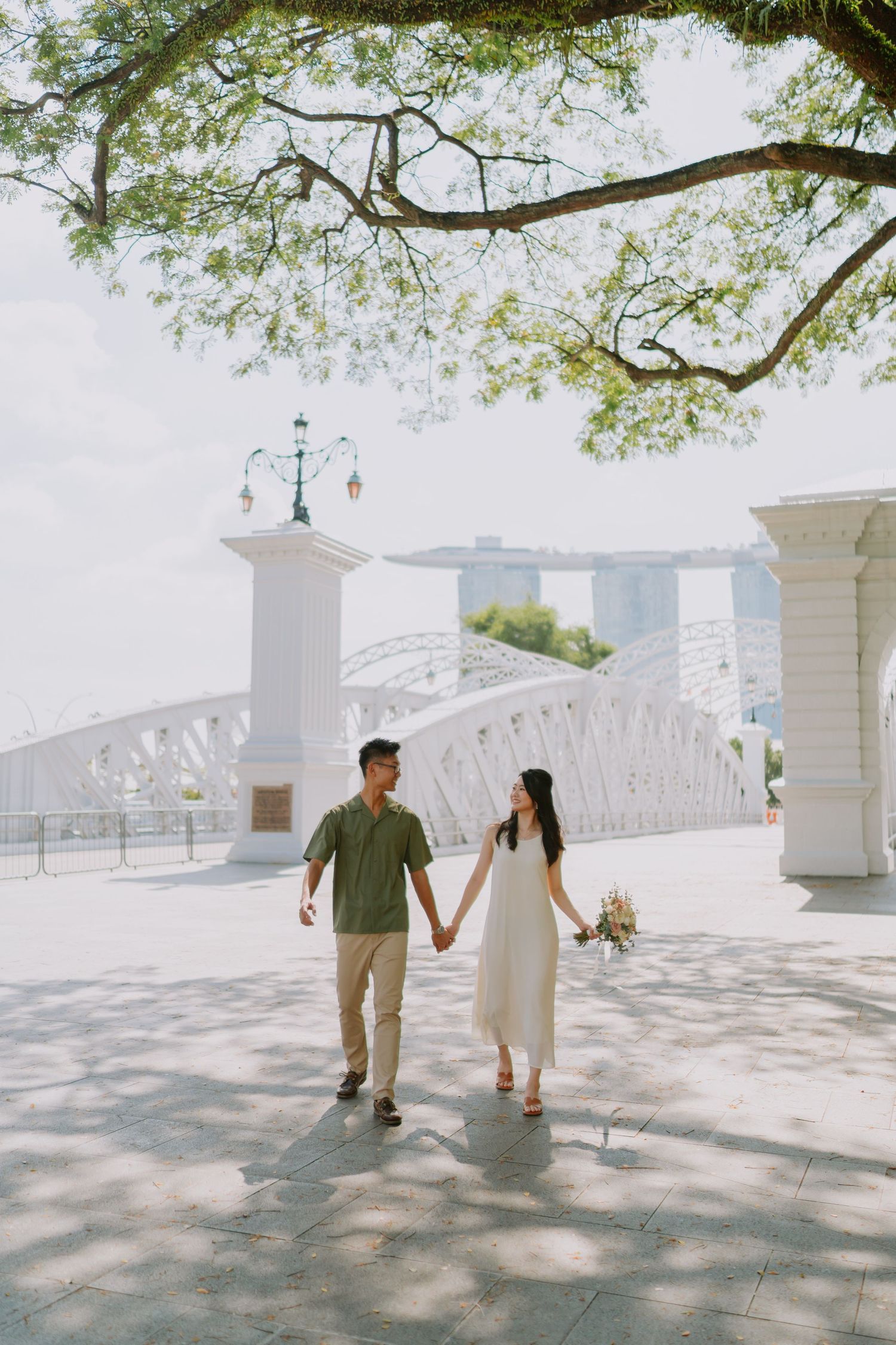 Couple holding hands walking near white bridge with Marina Bay Sands backdrop.