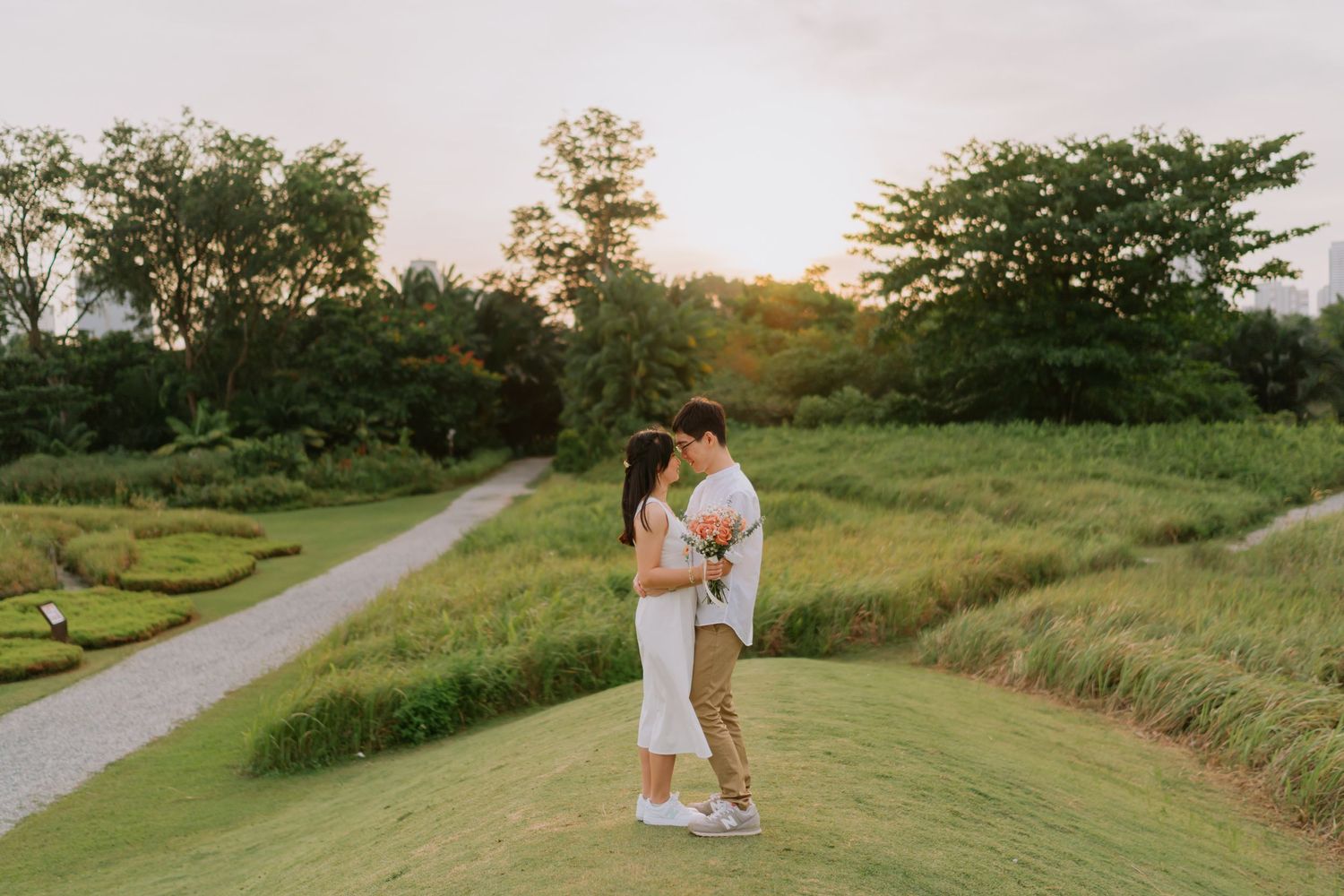 A couple shares a kiss on a scenic green hillside at sunset surrounded by lush trees and gardens.