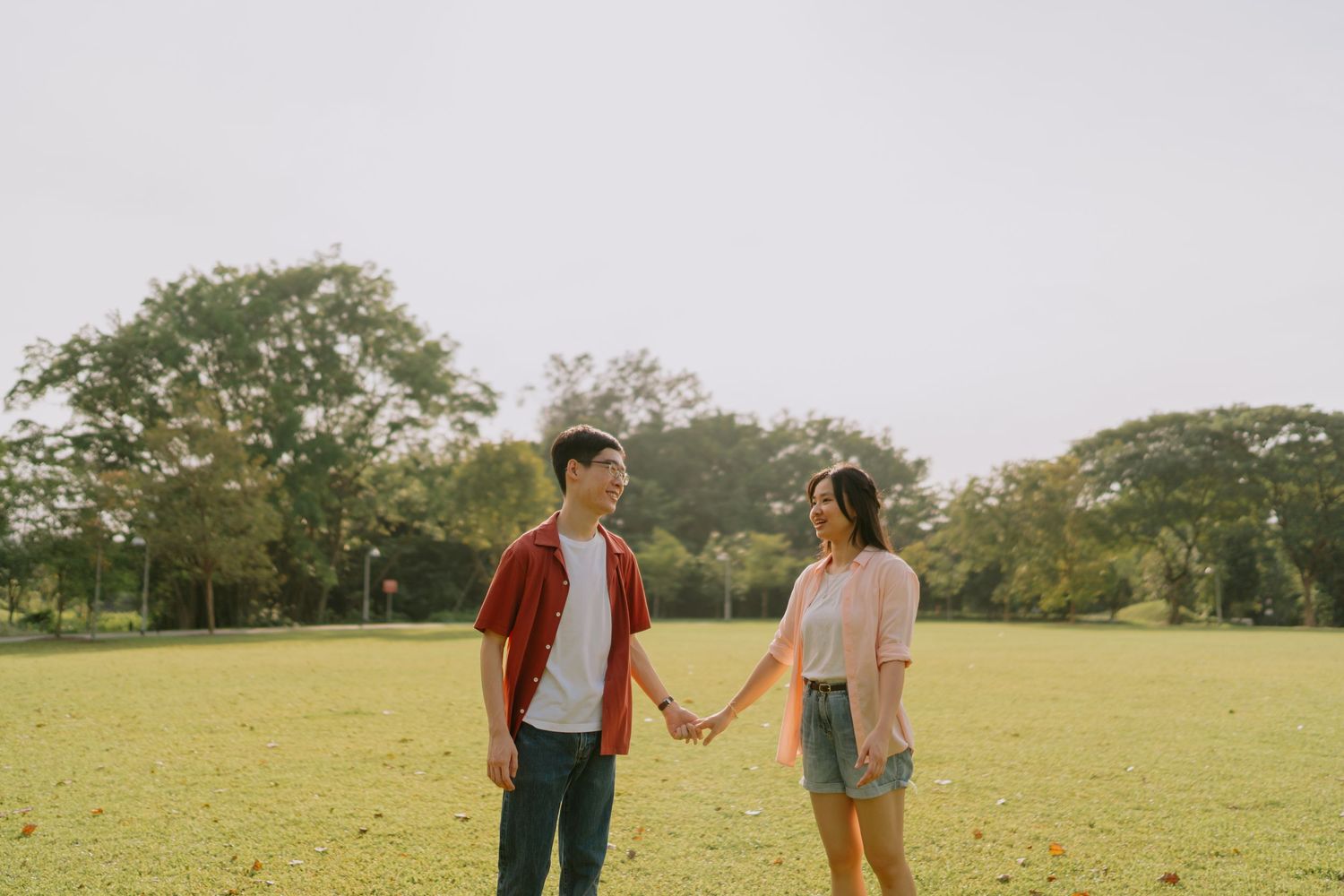 A young couple holds hands and gazes at each other in a lush green park on a sunny day.