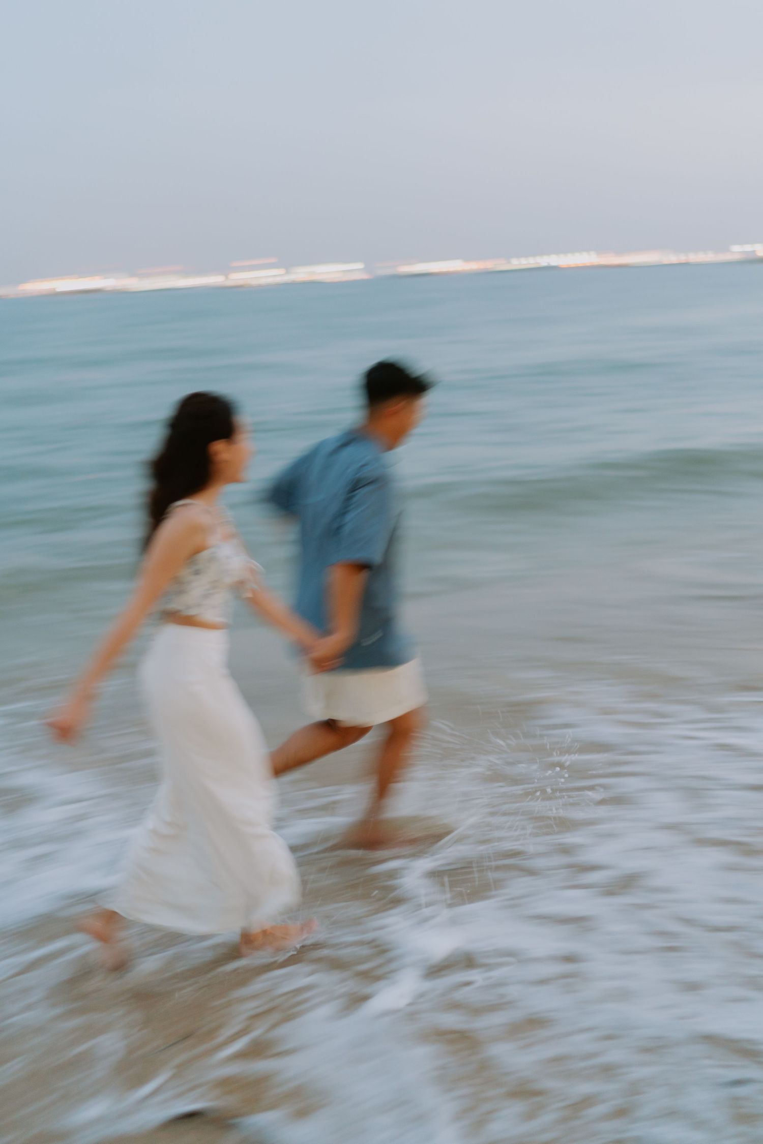 Blurred motion photo of a couple holding hands and running through beach waves at sunset.