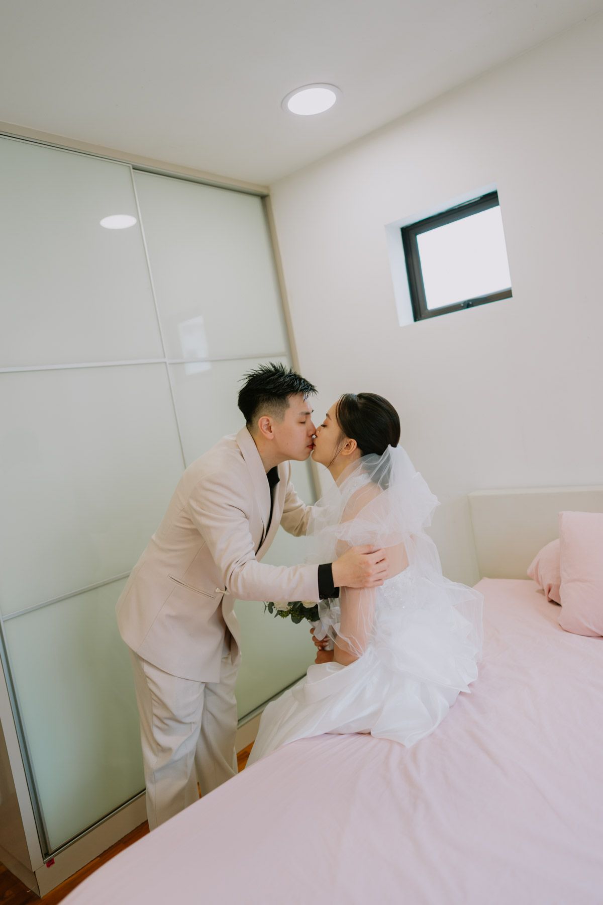 A bride and groom share a romantic kiss in a bright bedroom, with the bride wearing a white gown and veil.
