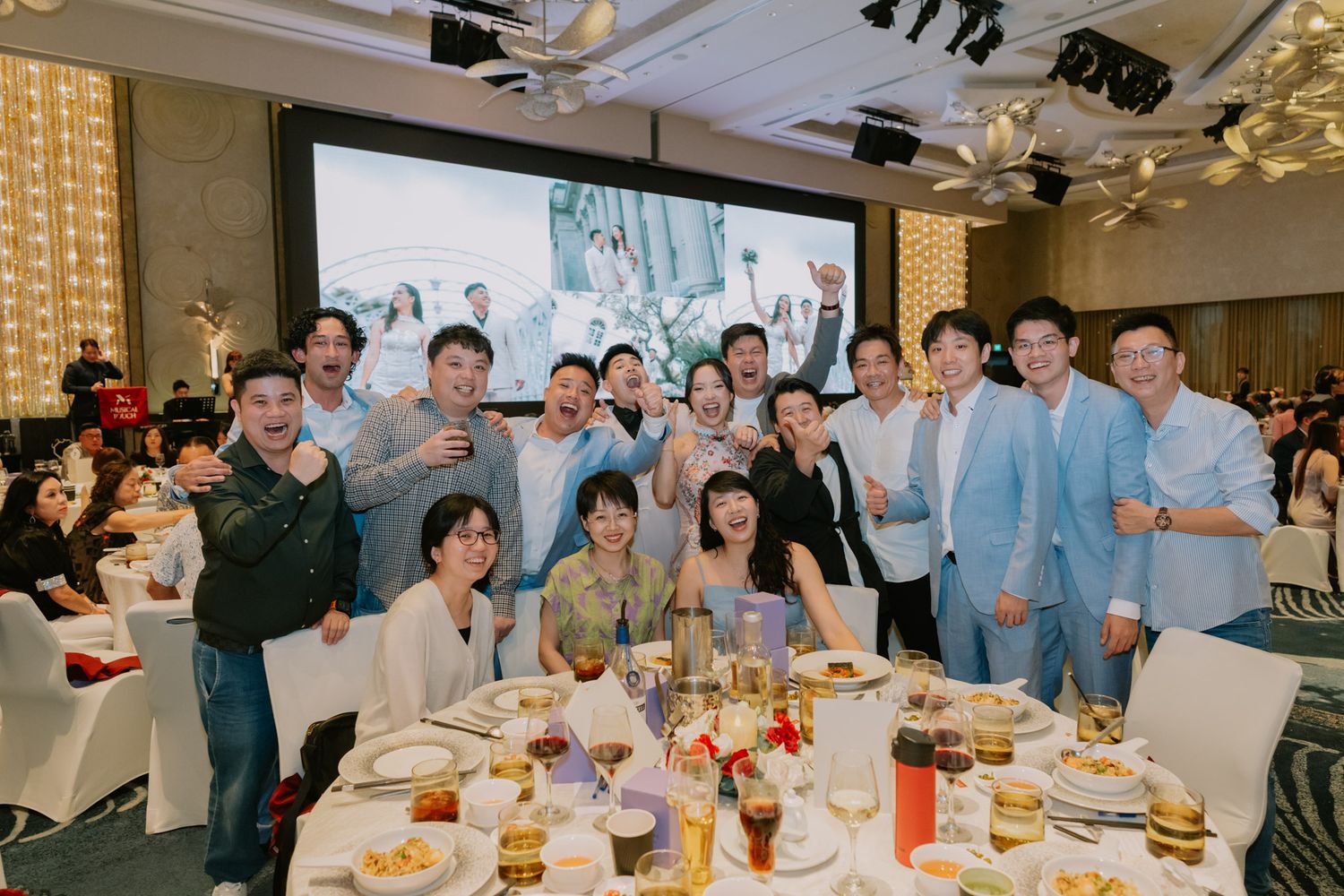 Group of colleagues posing together at a formal banquet dinner event in a ballroom with a large screen backdrop.