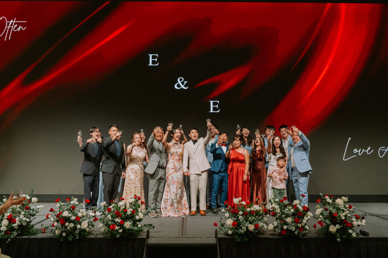 Wedding party celebrates on stage under E&E monogram with red floral decor and dramatic backdrop.