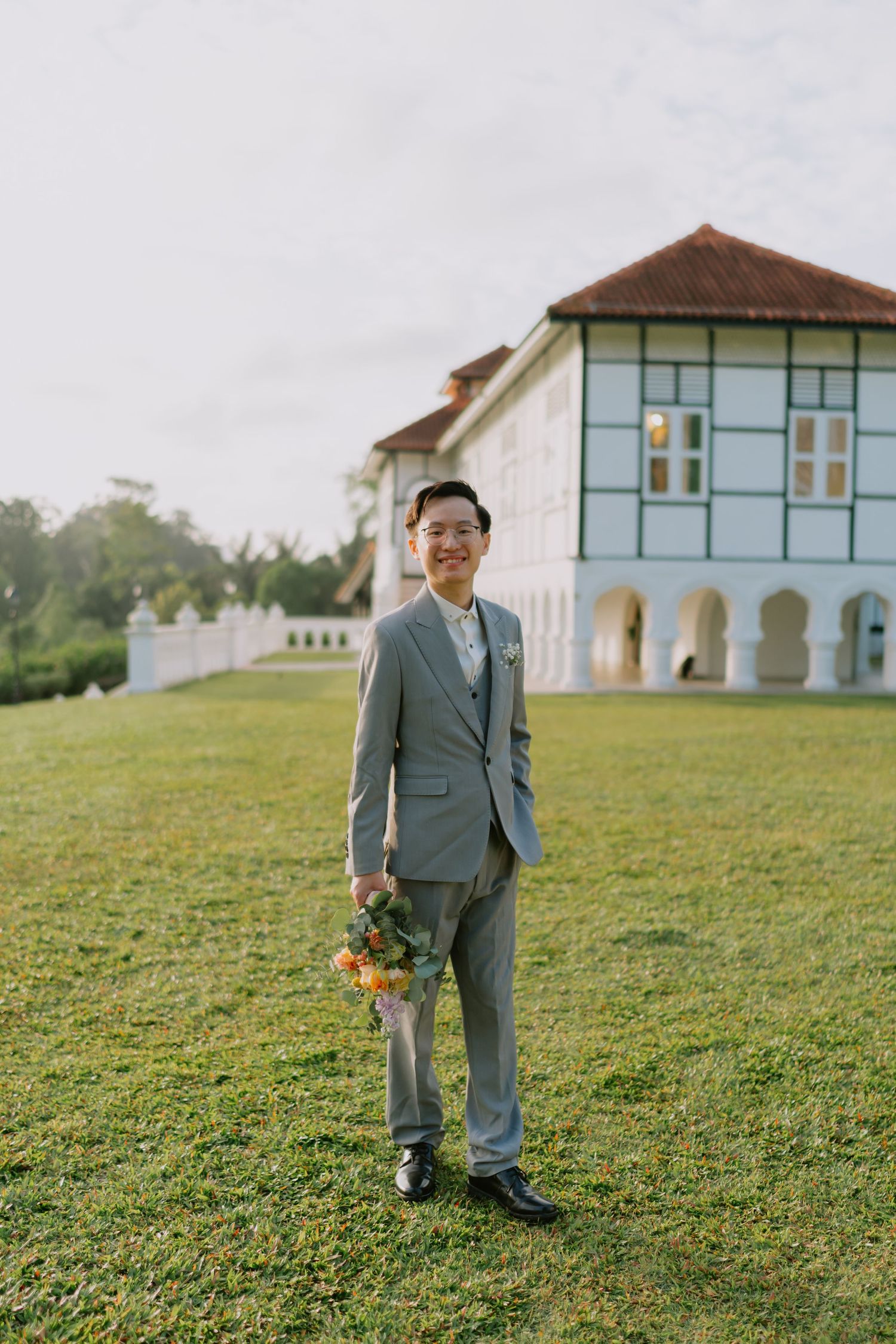 Smiling Asian groom in gray three-piece suit holds bouquet on green lawn before colonial building.