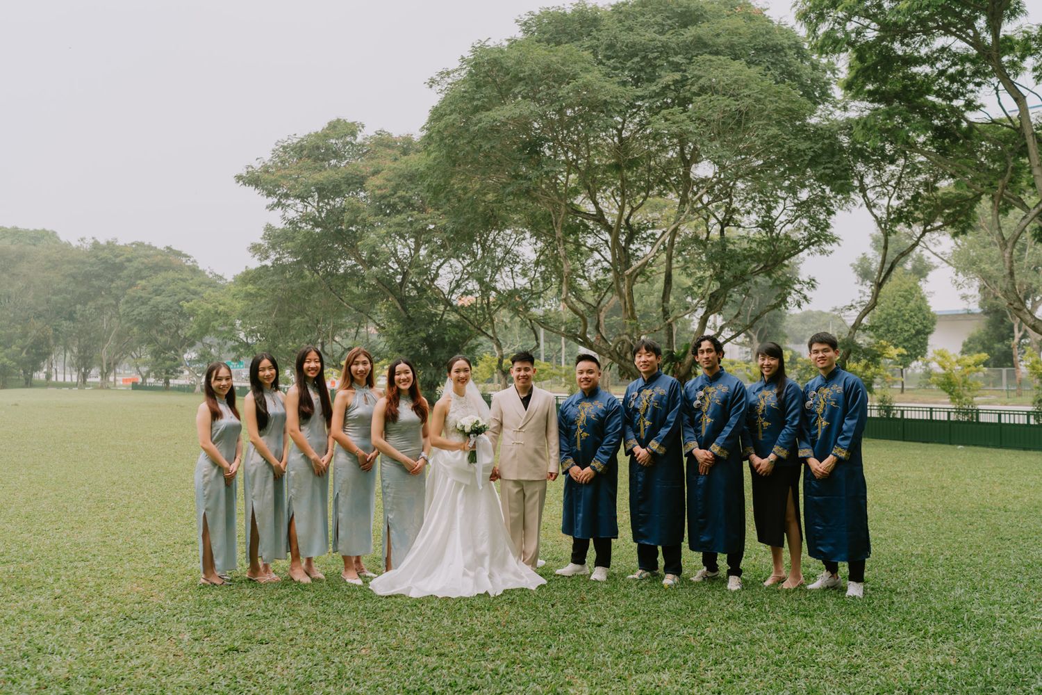 Wedding party poses on green lawn, bride in white gown, groom in tan suit, bridesmaids in sage dresses, groomsmen in navy suits.