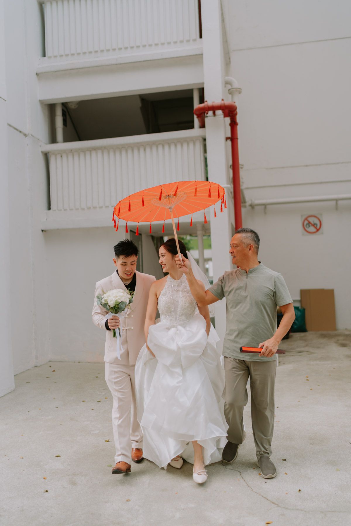 Bride in white gown walks with groom and elder holding red traditional umbrella outdoors at wedding ceremony.