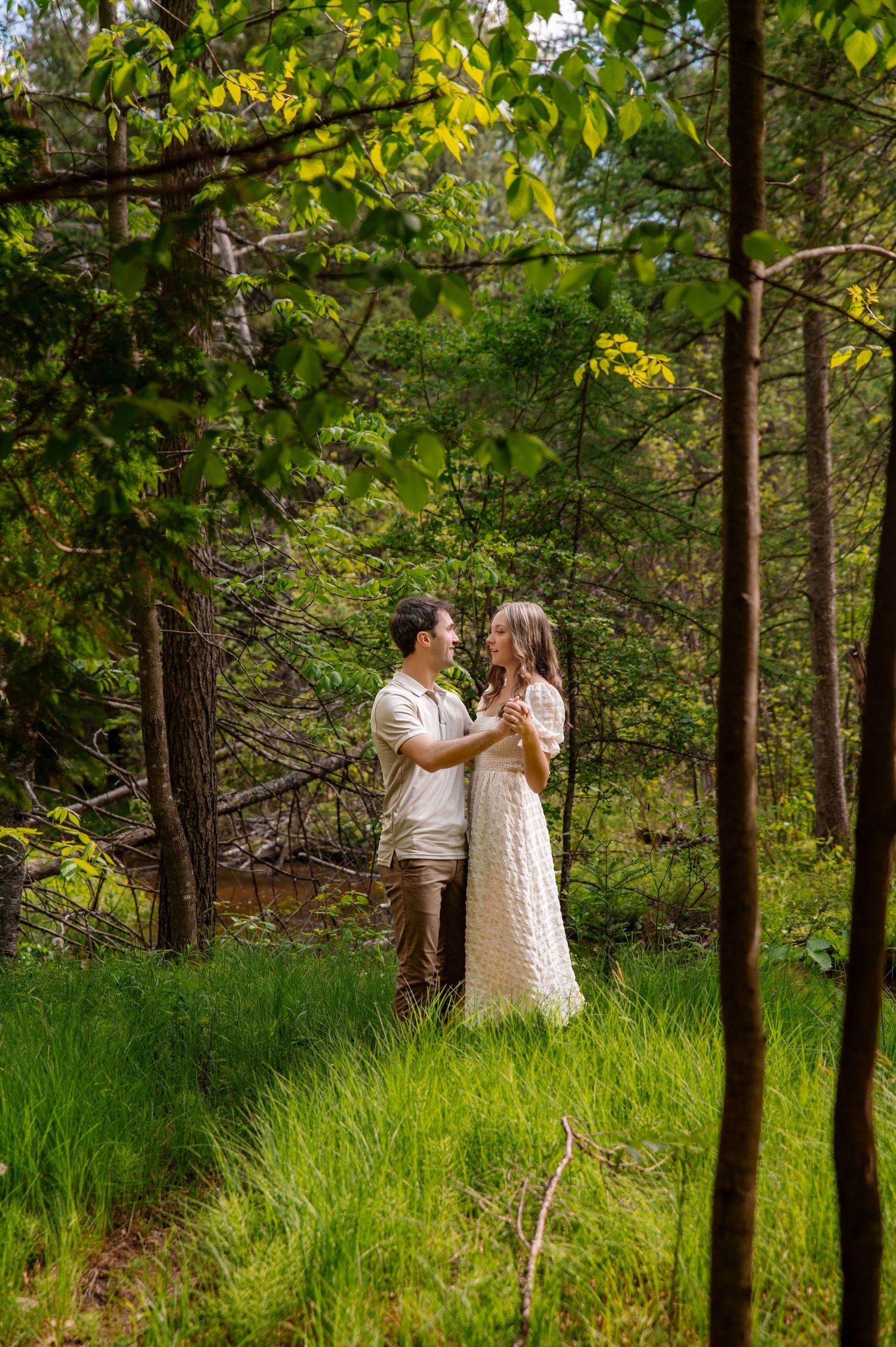 Romantic couple embraces in a lush green forest clearing wearing coordinated light-colored outfits.