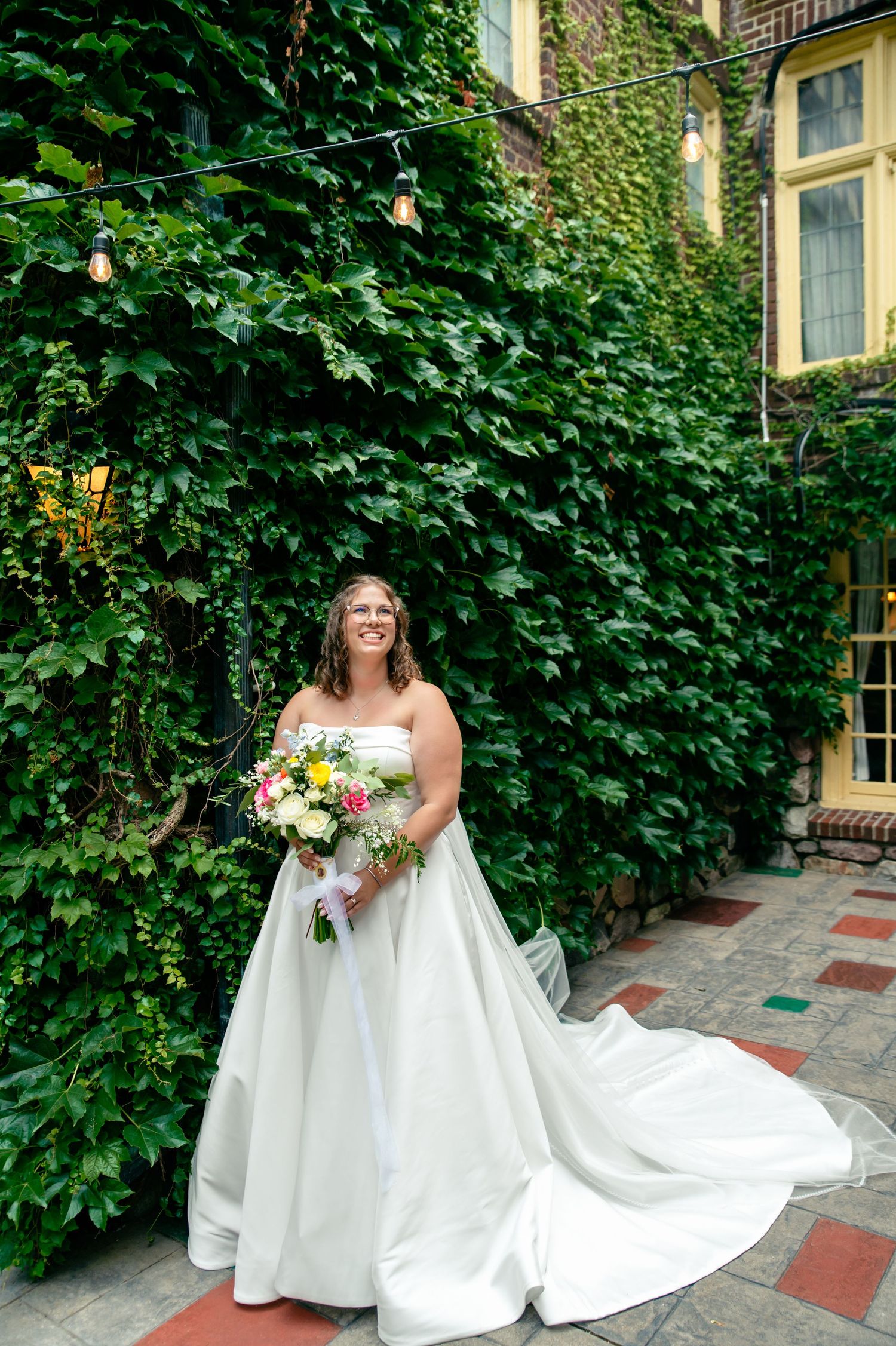 A bride in a flowing white wedding gown holds a colorful bouquet while standing in a lush garden courtyard.