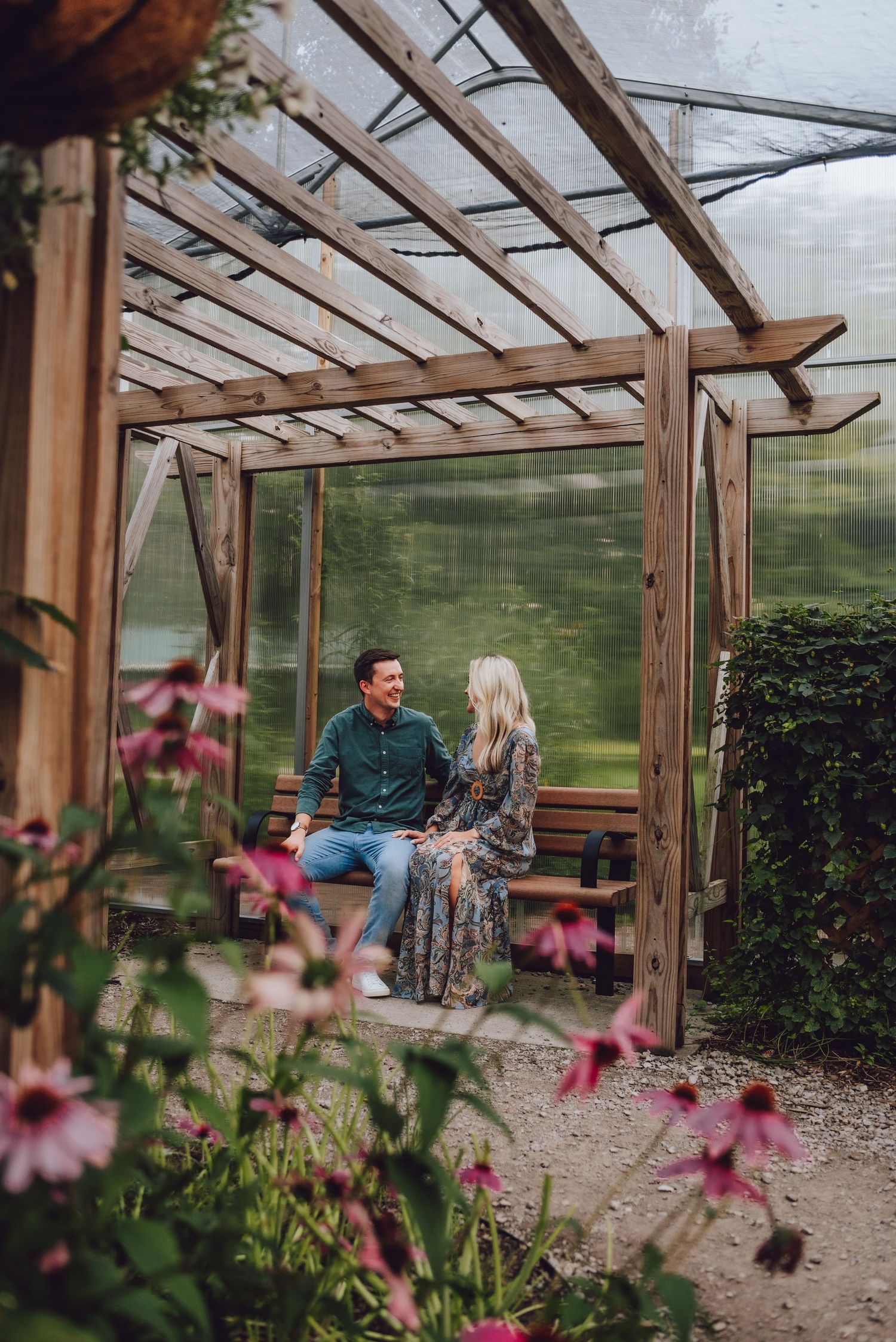 A couple sits on a wooden swing beneath a rustic pergola surrounded by pink flowers.