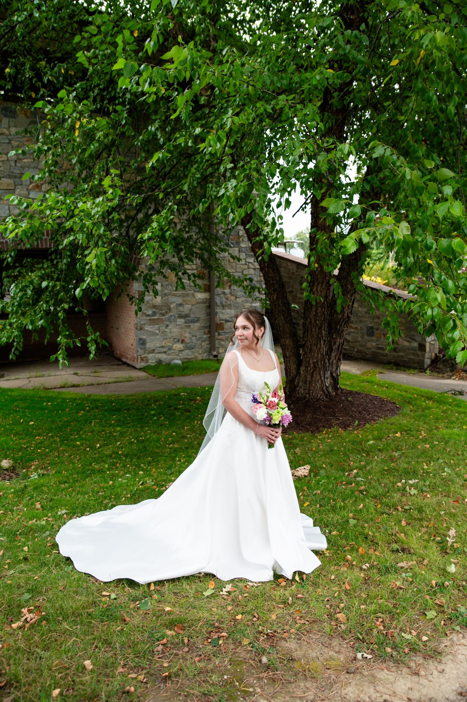 A person in a flowing white wedding dress stands on grass near a stone wall and tree holding a bouquet of flowers.