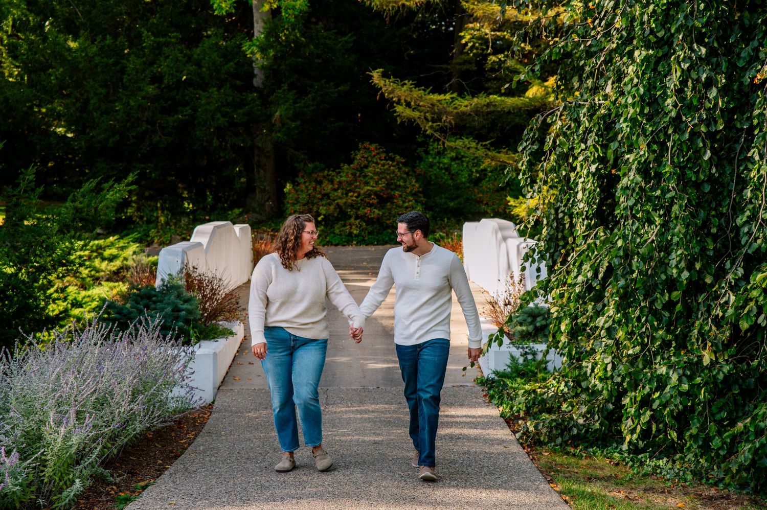 A couple in matching white sweaters walks along a garden path surrounded by greenery.