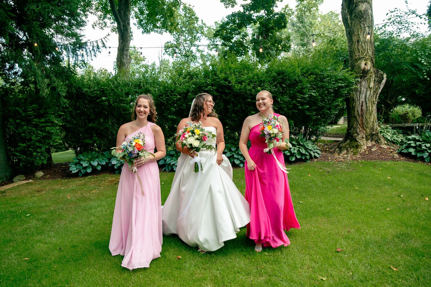 Three people in formal attire pose together on a grassy lawn during a summer wedding celebration.