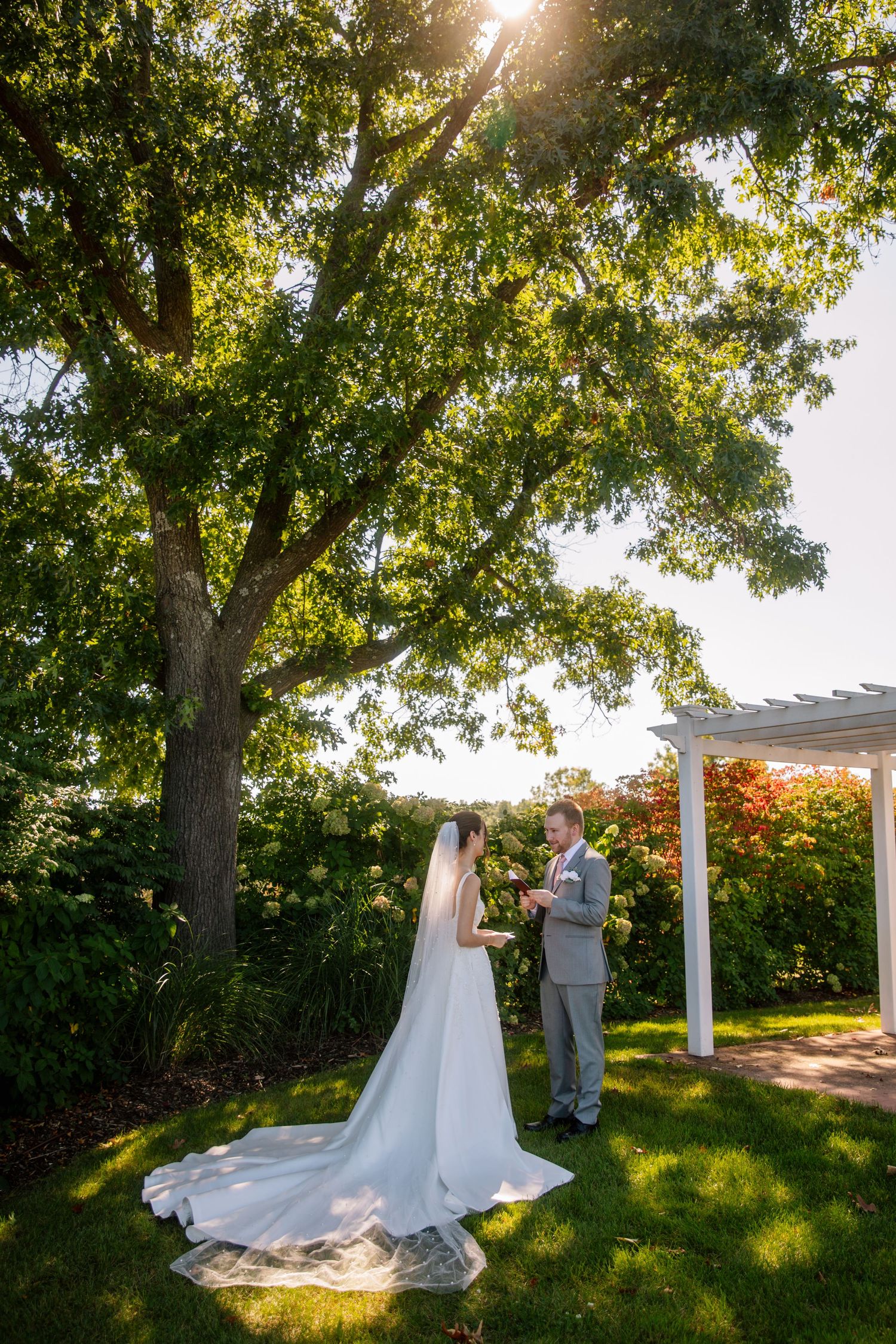 A wedding ceremony takes place under a white pergola surrounded by lush trees with sunlight streaming through the branches.