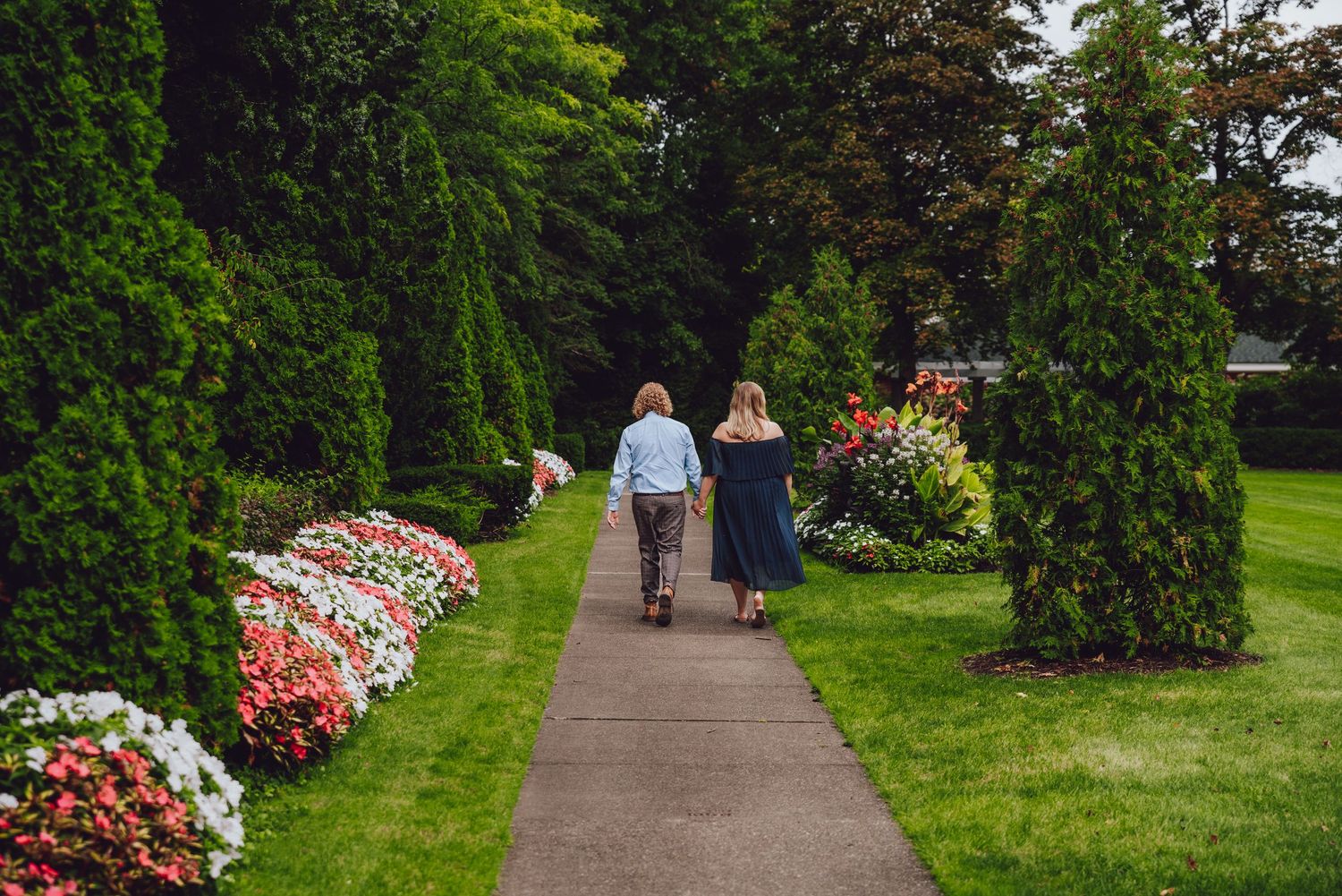 Couple walking along garden path bordered by colorful flowers and evergreen trees.