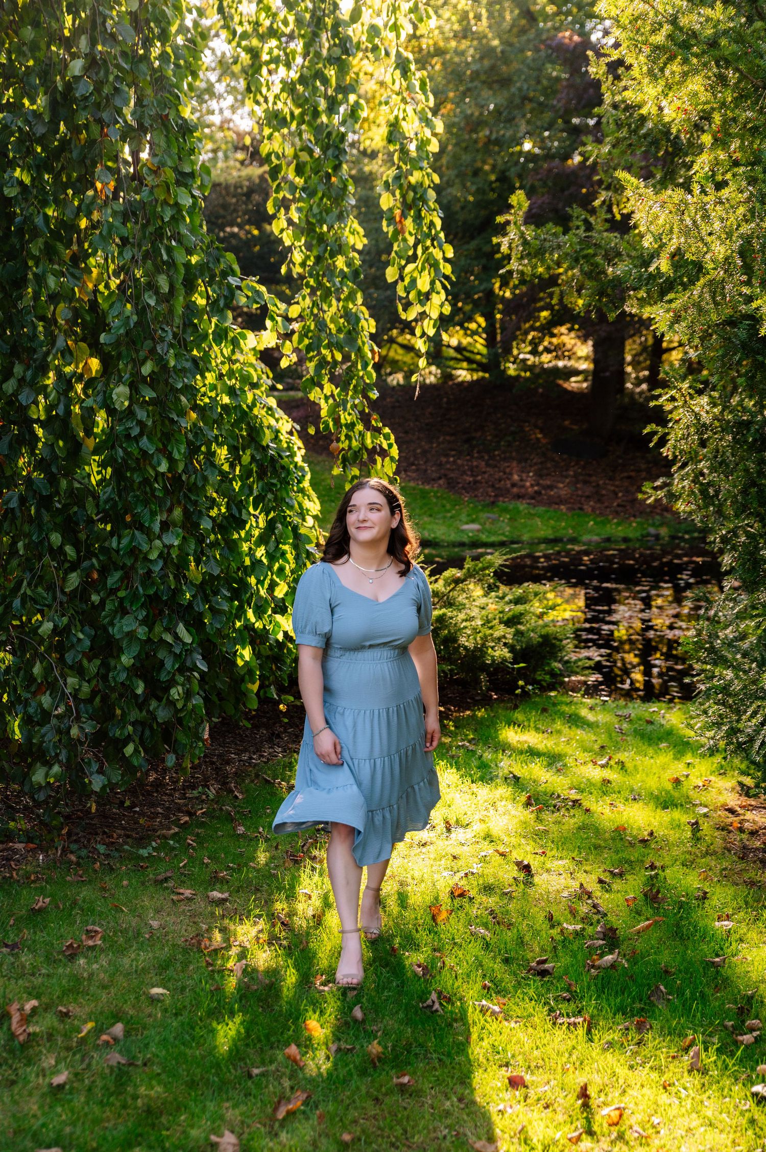 Woman in blue dress walking through sunlit garden with hanging vines and dappled light.