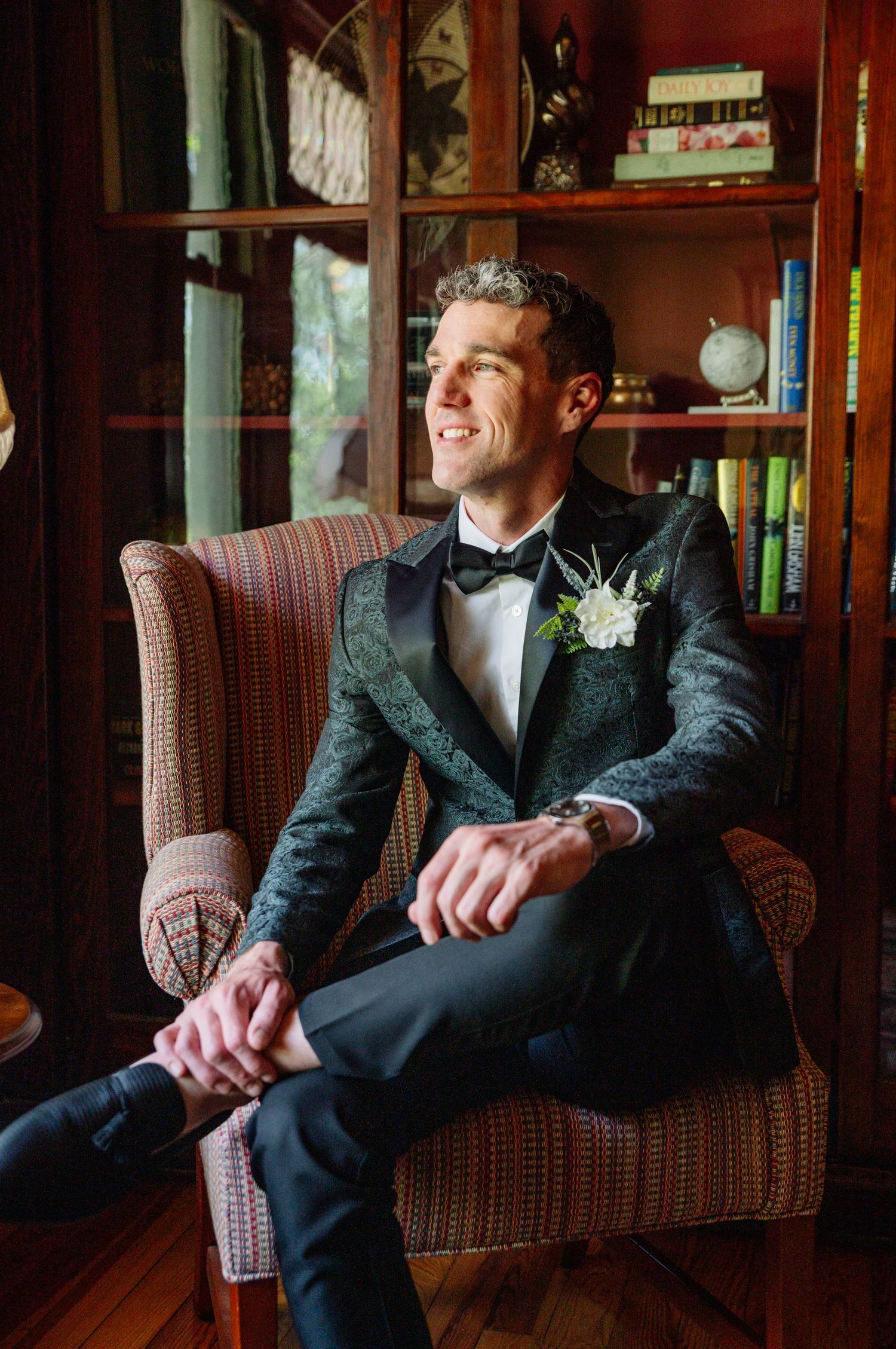 A groom in a black tuxedo and bowtie sits relaxed in a vintage armchair while smiling in a warmly lit room.