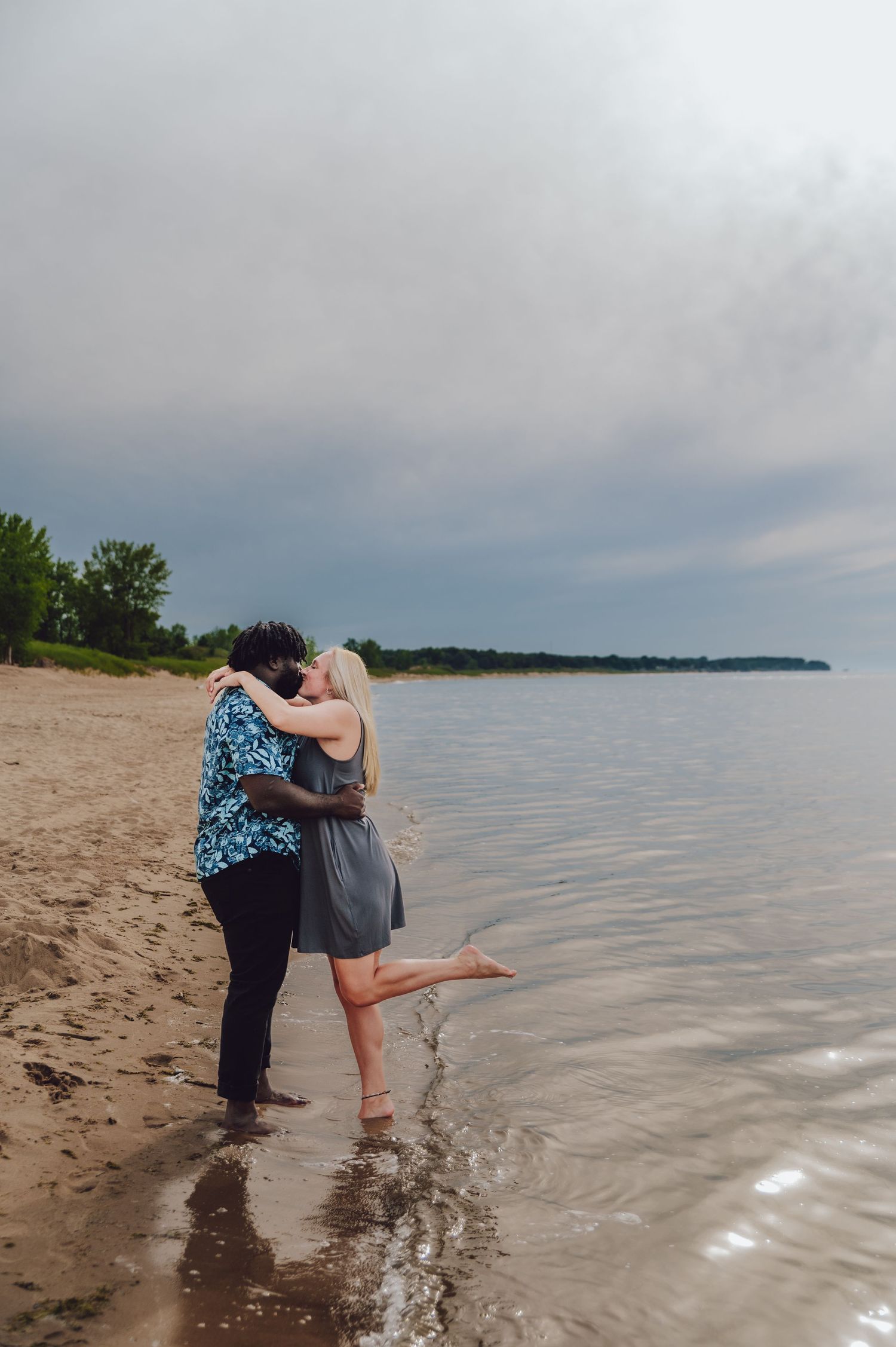 Couple sharing a romantic moment on a beach shoreline during overcast weather.