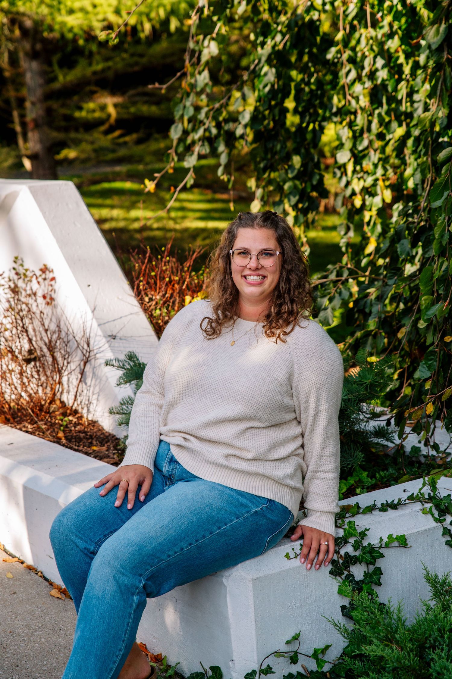 Person in cream sweater and jeans sits casually on white garden wall surrounded by greenery.