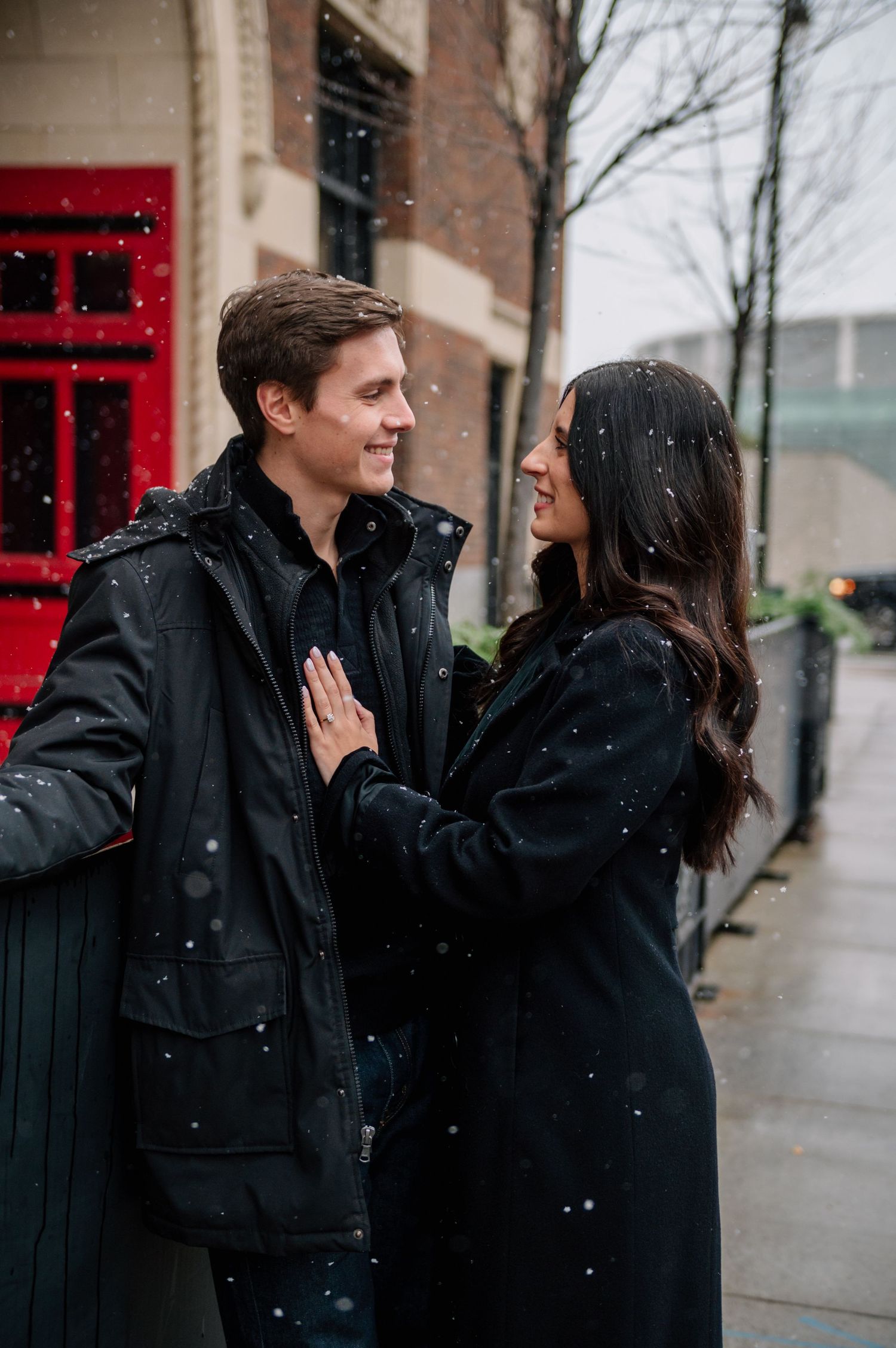 A romantic couple shares an intimate moment in the snow outside a building with a red door.