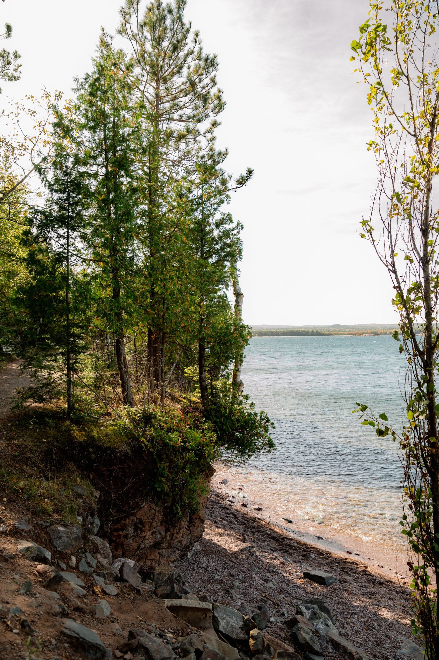 Rocky shoreline on a tranquil lake with trees and shrubs growing along the water's edge during daytime.