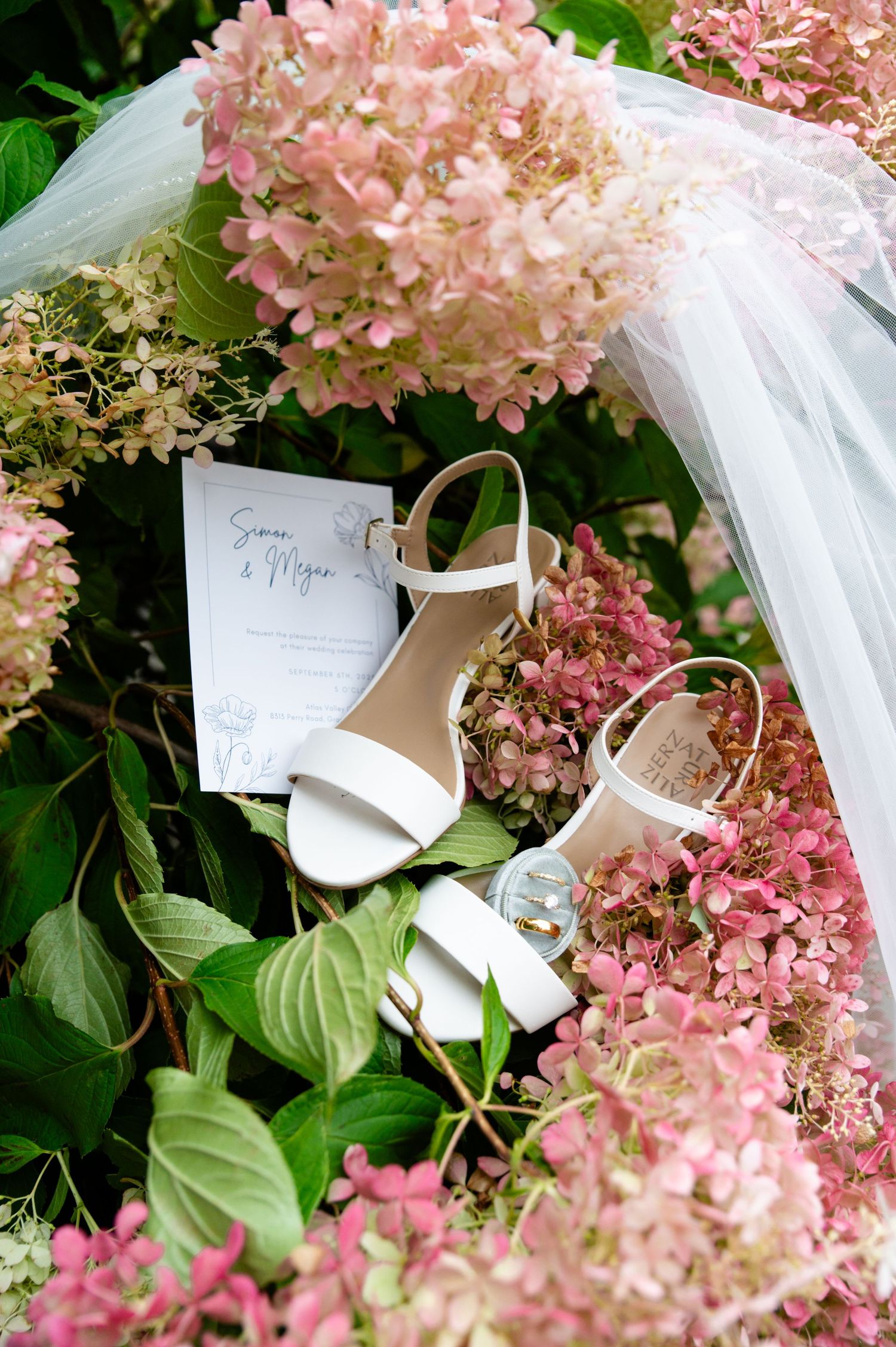 Pink hydrangea flowers with white wedding invitation cards nestled among green leaves in an outdoor garden setting.