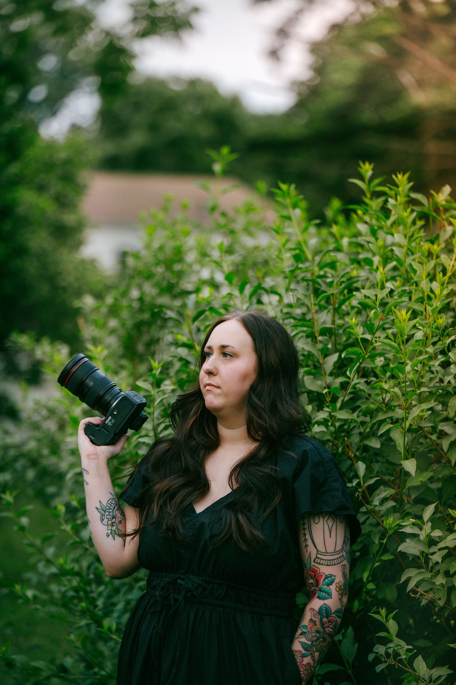 A photographer in a black dress stands among green foliage taking pictures with a professional camera in nature.