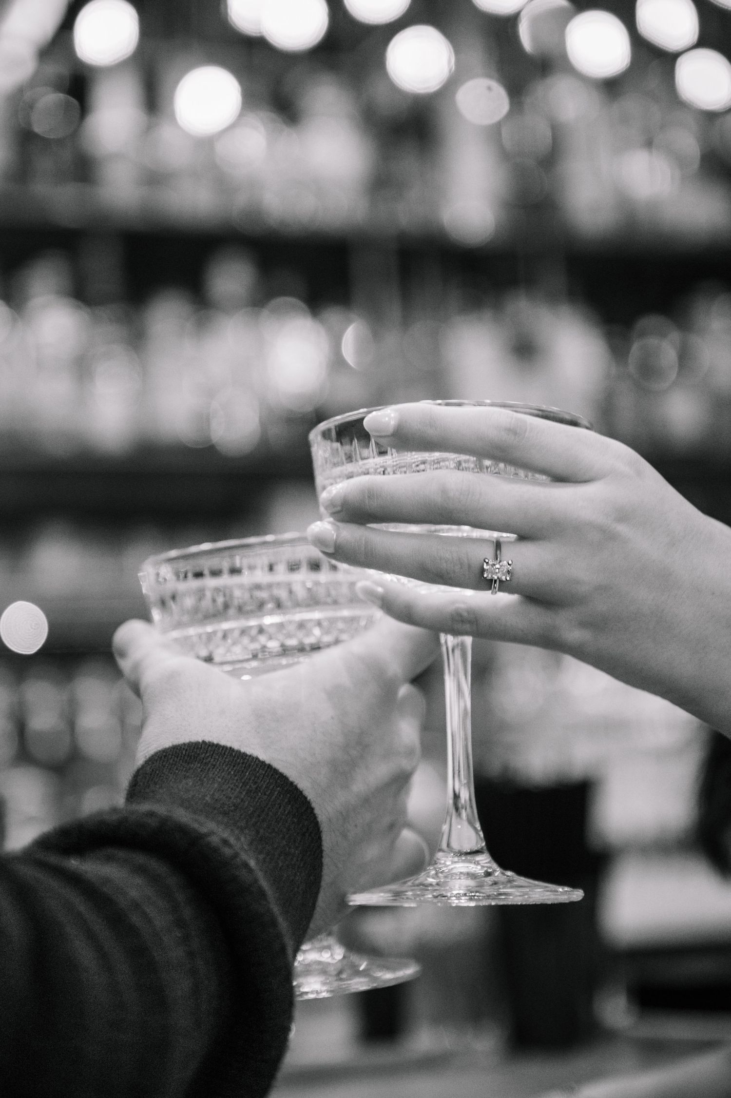 Two hands toasting champagne glasses at a bar, with an engagement ring visible.