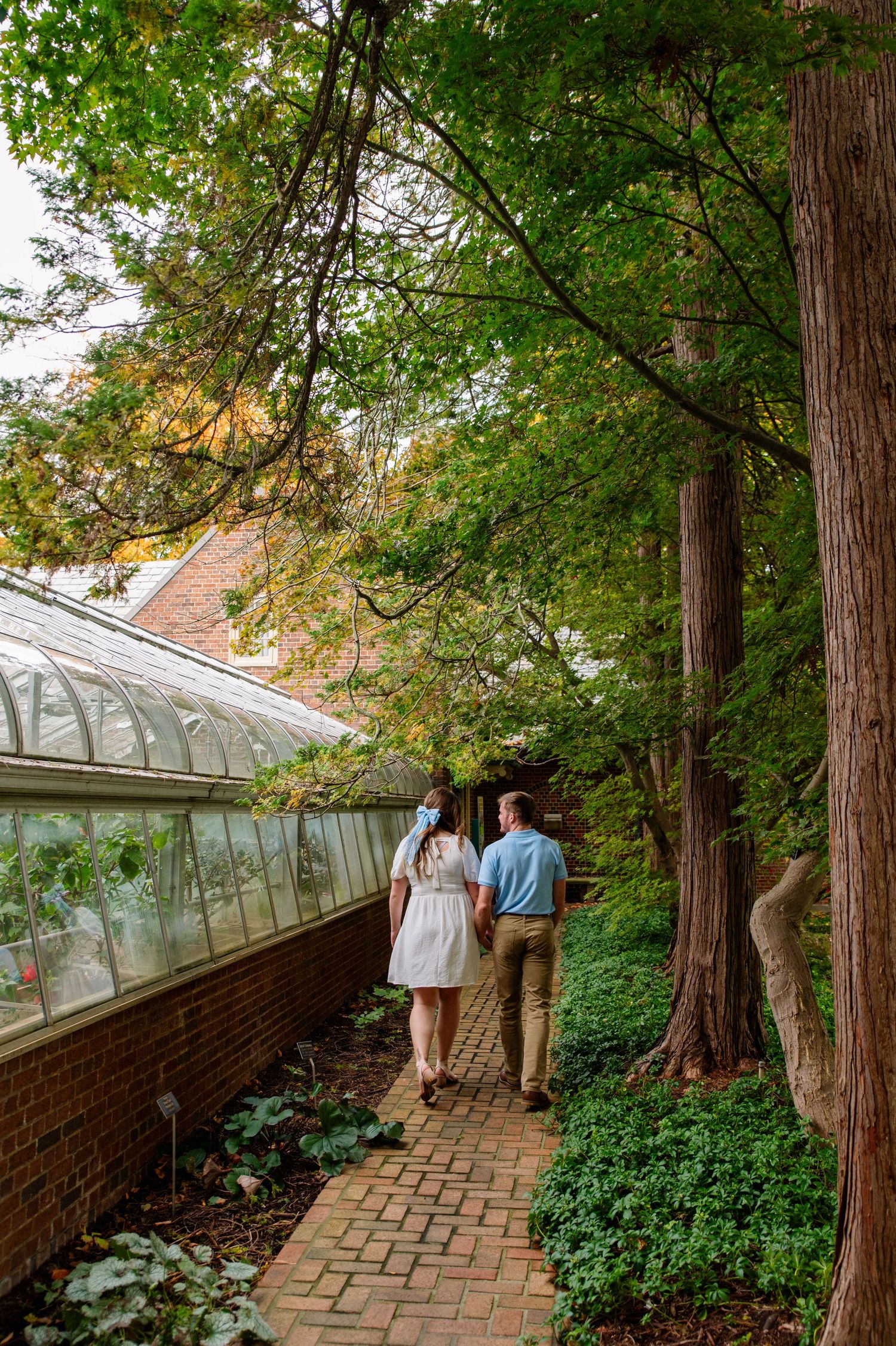 Couple walks hand in hand along brick path next to greenhouse during engagement session.