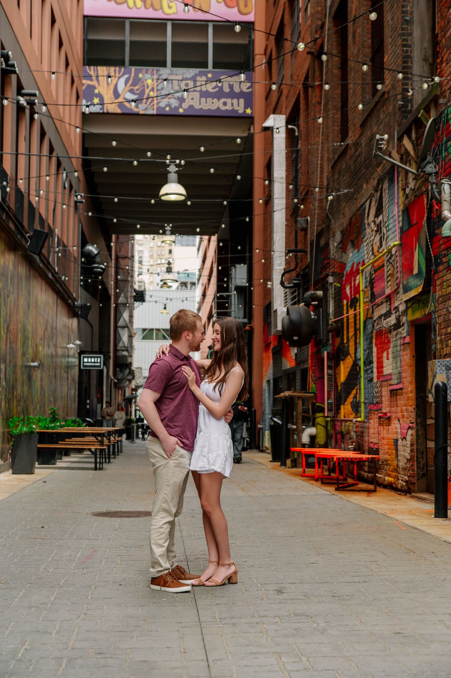 A couple embraces in a trendy industrial alley with exposed brick walls and string lights.