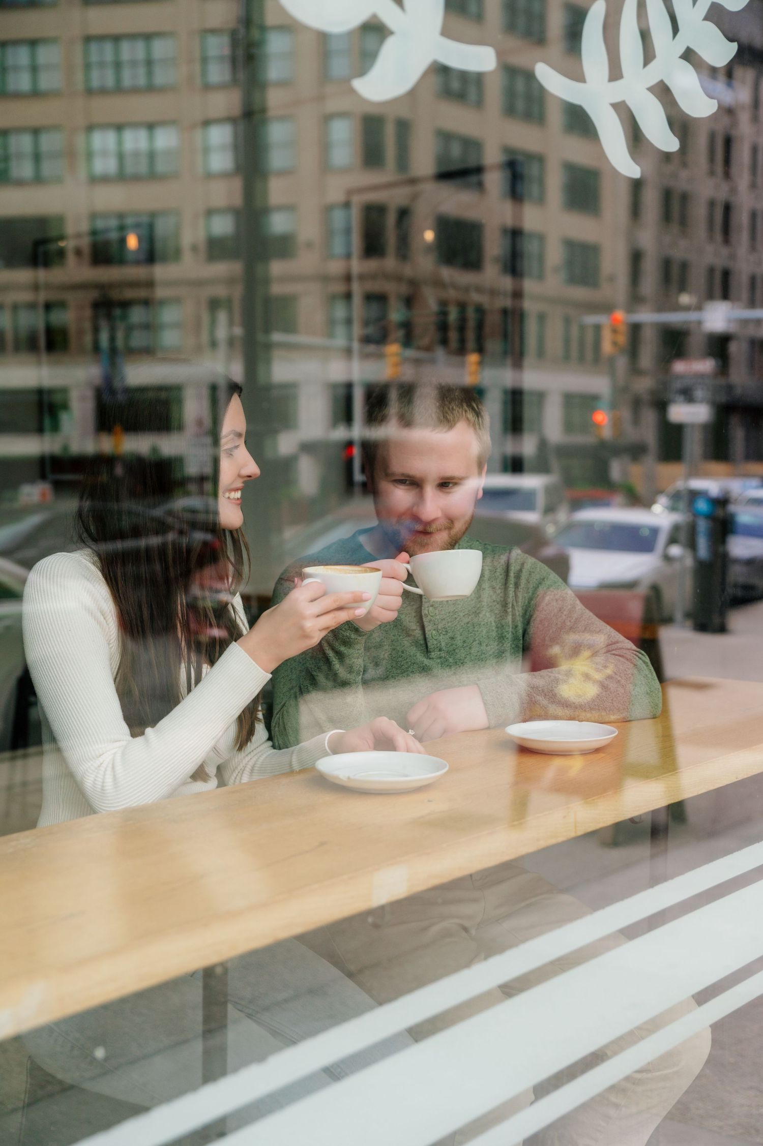Two people enjoying coffee at a cafe table viewed through a window.