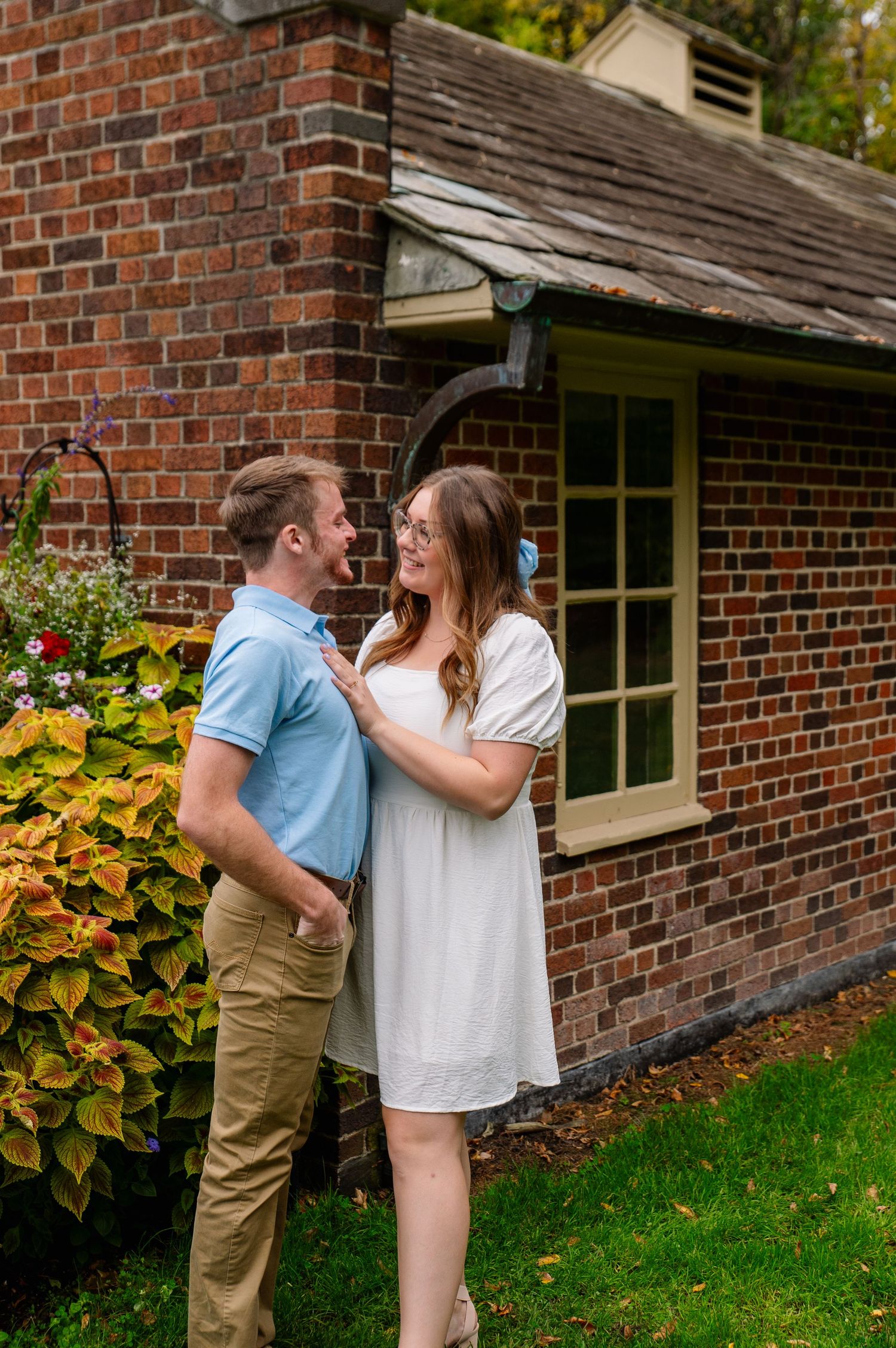 Couple shares intimate moment against brick wall with colorful garden backdrop.