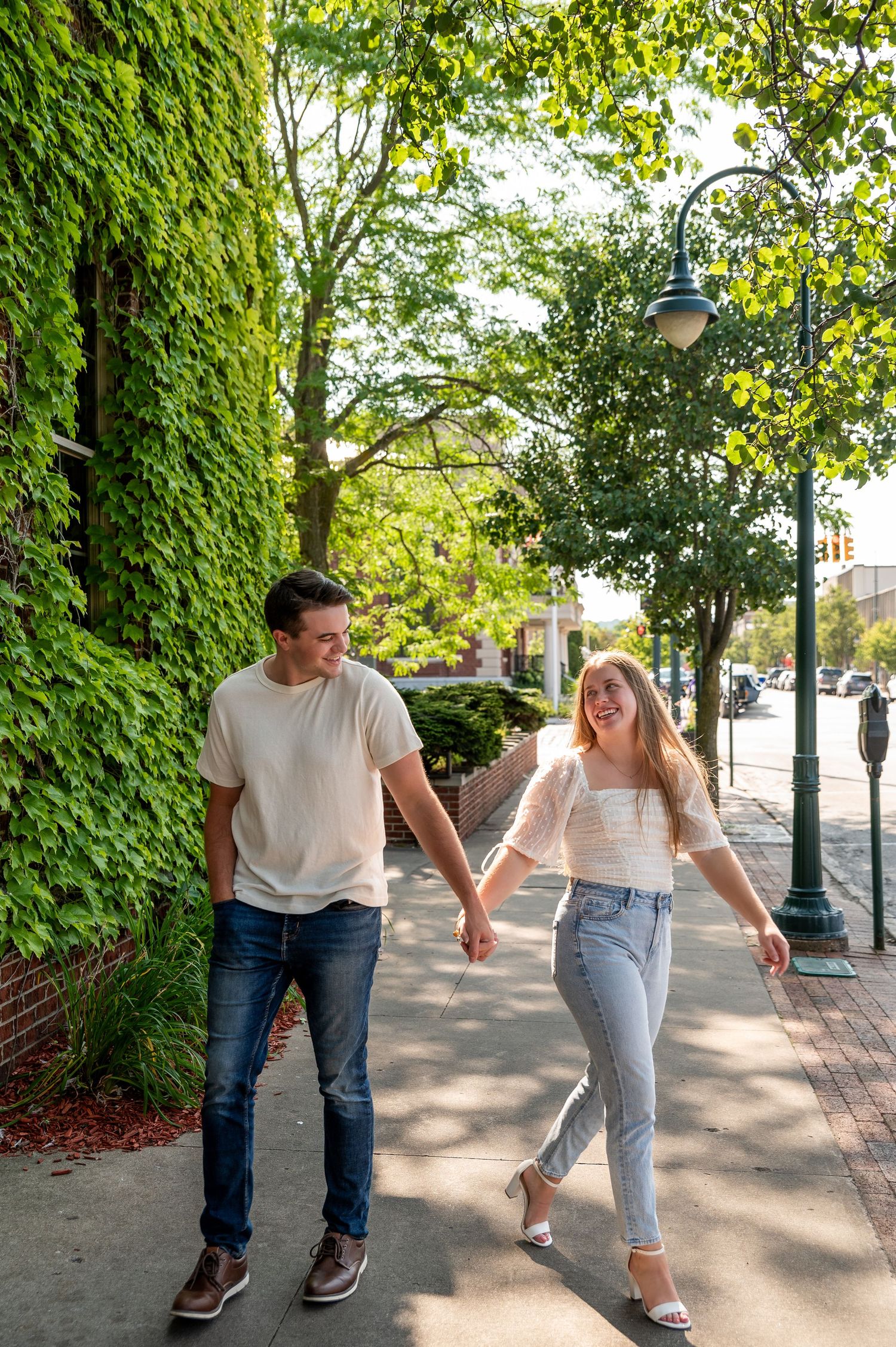 A couple walks hand in hand along a tree-lined sidewalk on a sunny day.