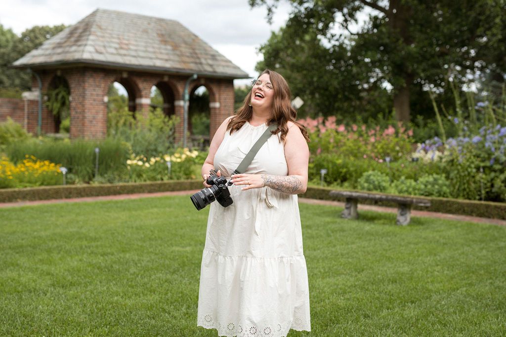Professional photographer in a white lace dress walks on a grassy hillside while carrying camera equipment.