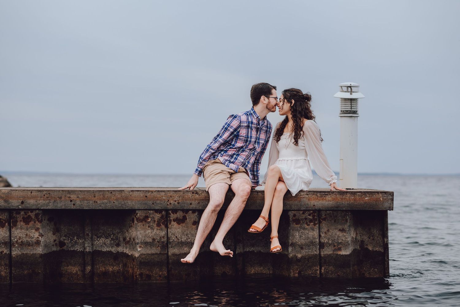 A romantic couple sits barefoot on a pier by a lighthouse over calm waters at dusk.