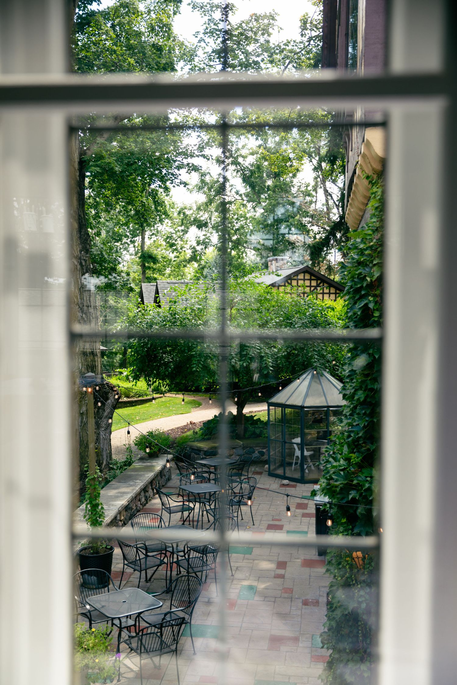 View through a window of an outdoor patio with metal tables and chairs under trees, featuring a decorative gazebo.