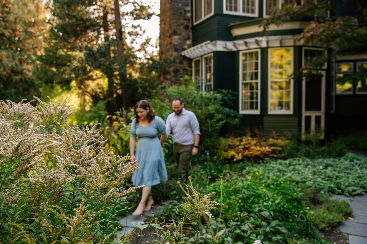 Couple walking through lush garden path beside vintage house at sunset.