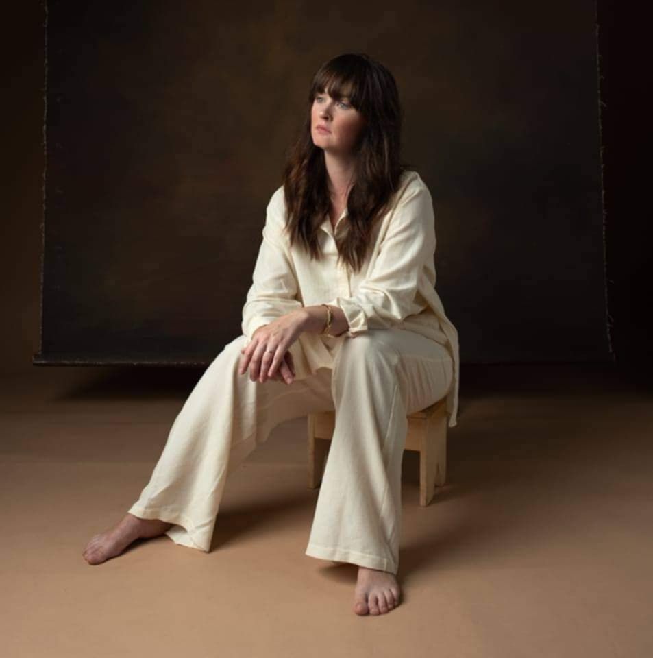 Professional portrait of a person in cream colored outfit sitting barefoot on chair against dark backdrop.