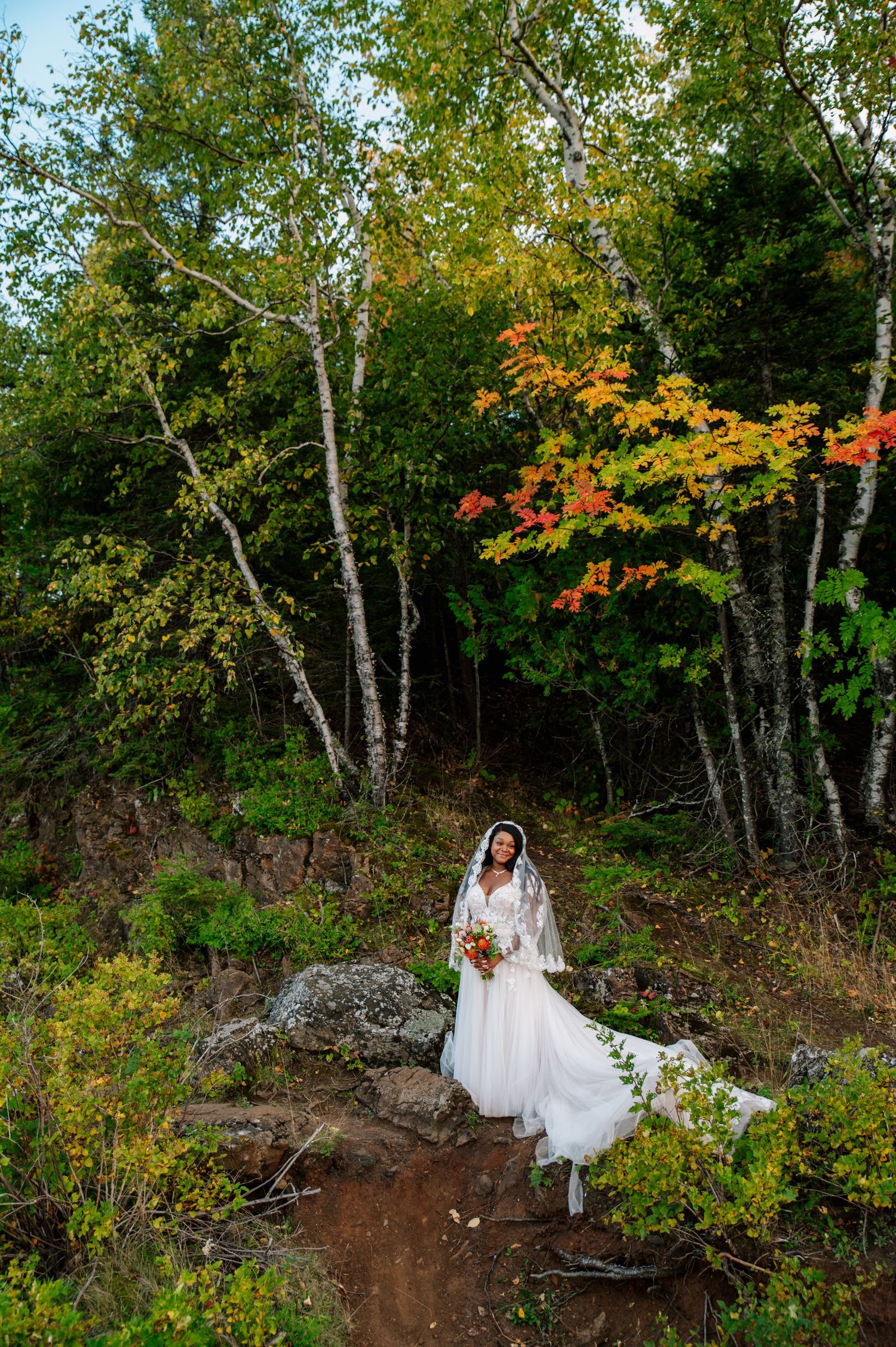 A person in a white wedding dress poses in a scenic autumn forest setting with birch trees and fall foliage.
